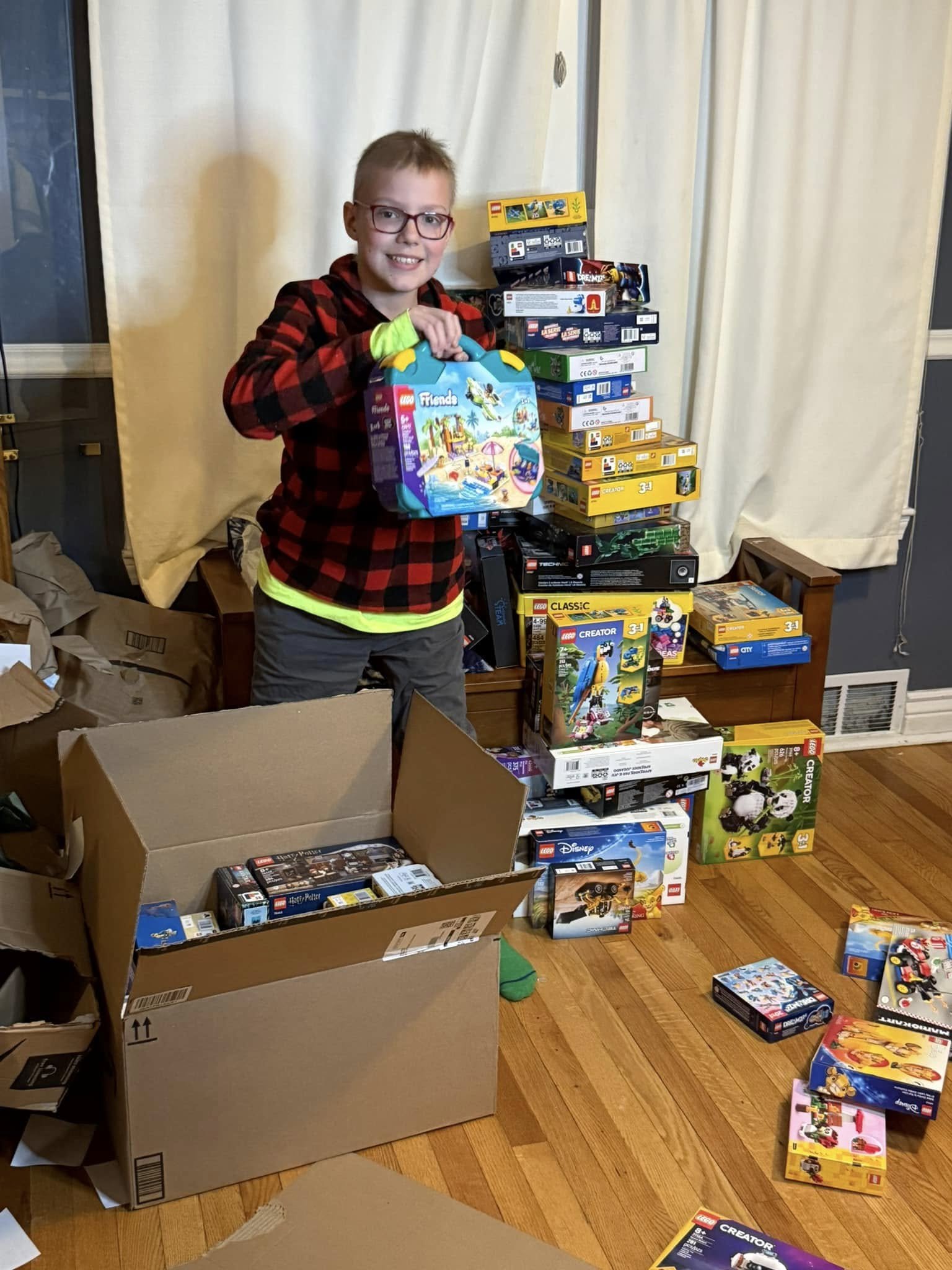 A young boy with glasses and a red and black checkered jacket holding a LEGO Friends set, standing in front of a large collection of LEGO boxes on the floor and a tower of stacked LEGO sets behind him.
