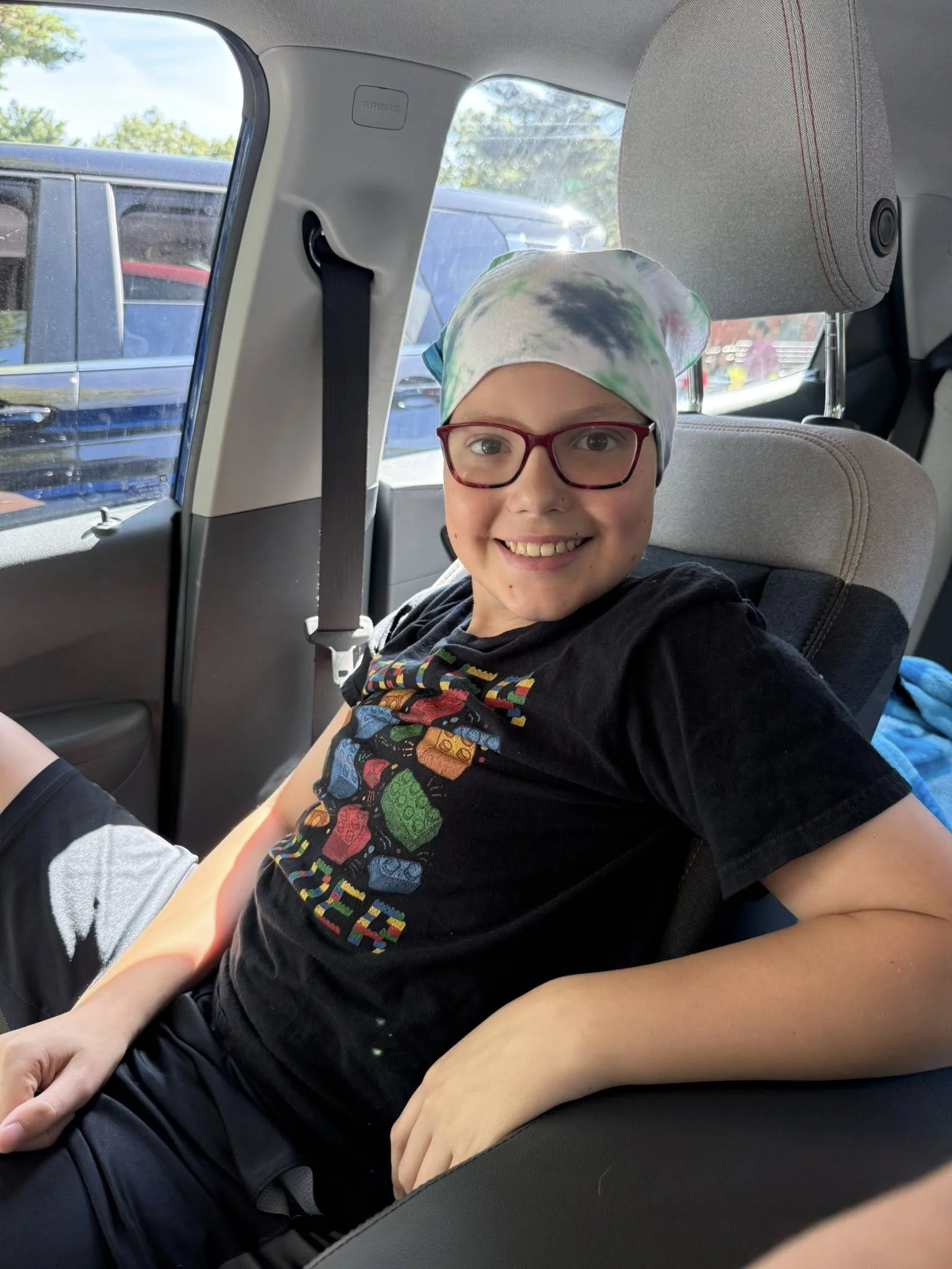 Young boy with glasses, smiling, sitting in a car seat in the back seat of a vehicle, wearing a colorful shirt and a tie-dye headscarf.