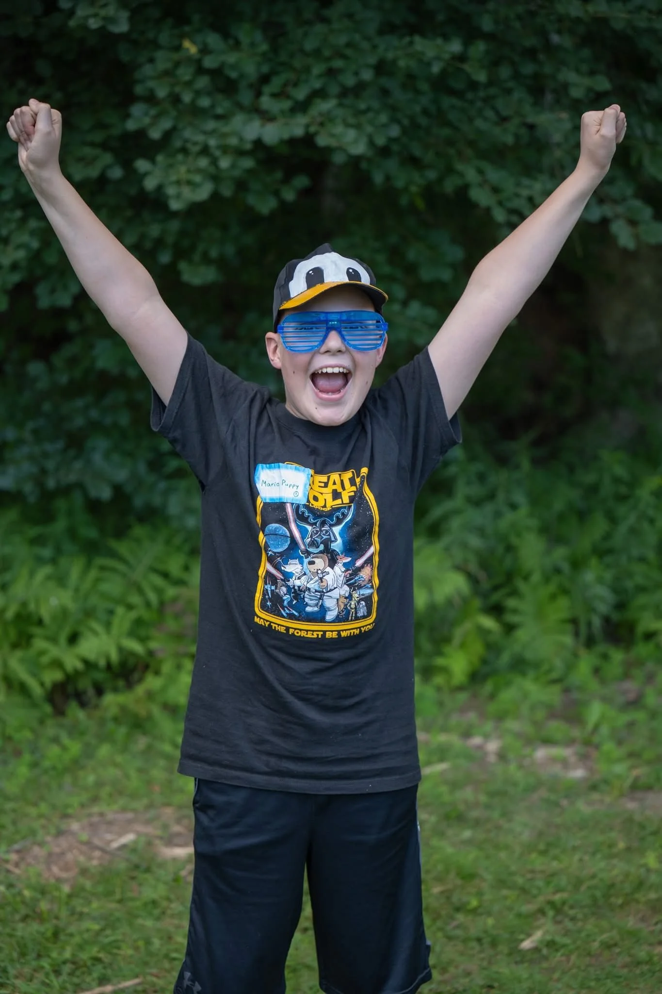 A boy celebrating outdoors with arms raised, wearing blue shutter shades, a black Star Wars t-shirt, and a penguin hat.