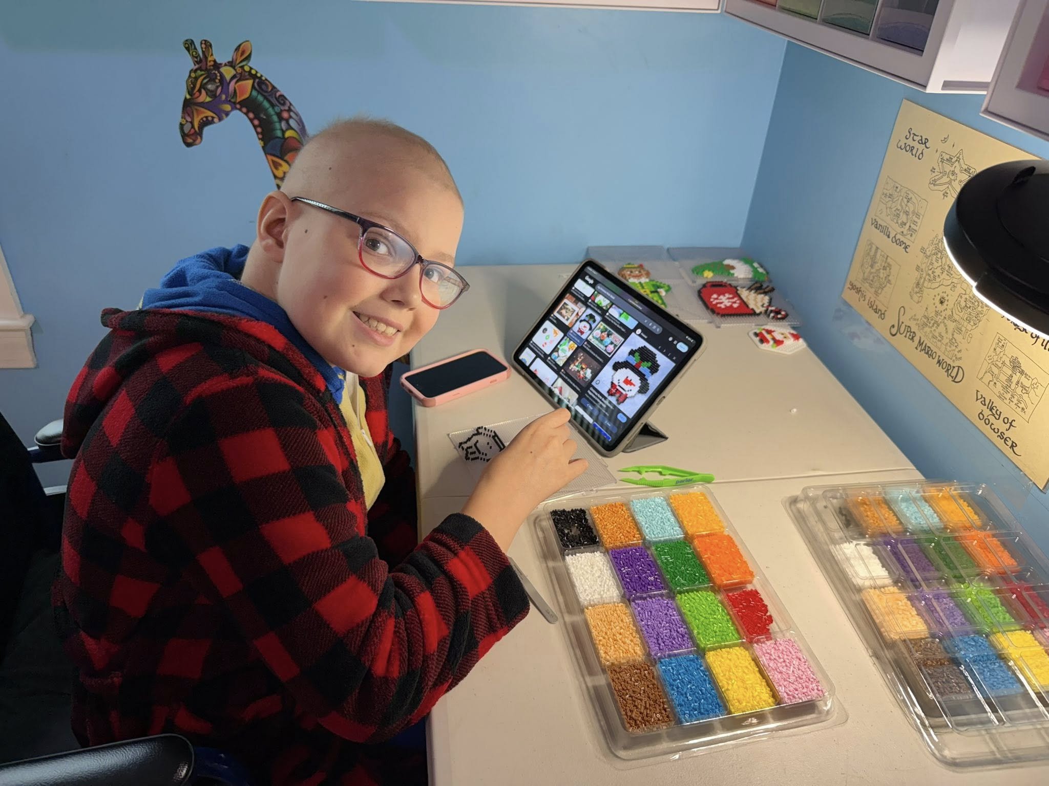 A young boy with glasses sitting at a desk, smiling at the camera, working on a tablet with pixel art designs on the screen. The desk has containers loaded with colored beads, a smartphone, and green craft scissors. Behind him is a blue wall with a c