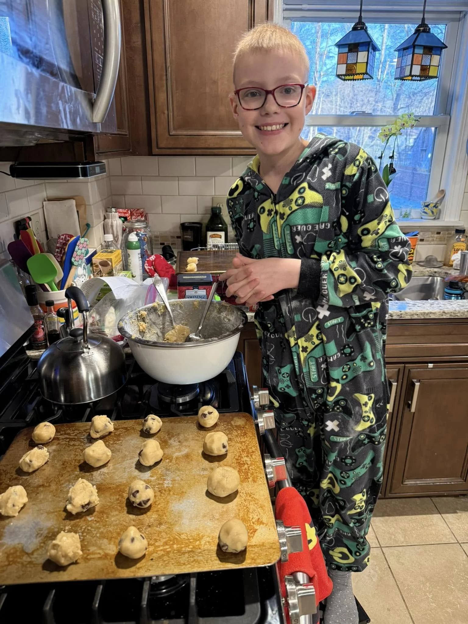 A smiling boy in colorful pajamas baking chocolate chip cookies in a kitchen. The boy is standing next to a baking sheet with cookie dough balls on it and preparing more cookies. There is a mixing bowl, a spoon, and various kitchen items on the count