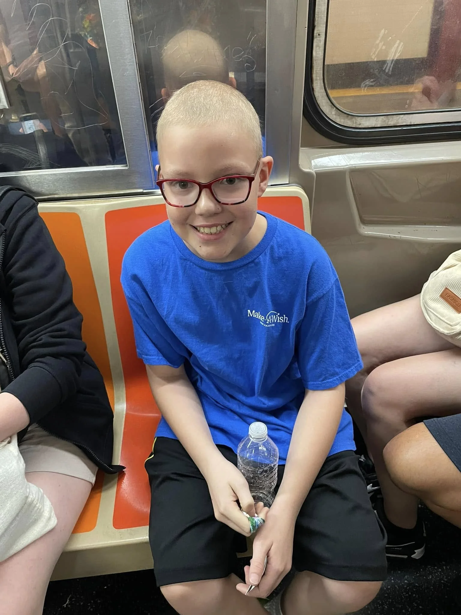 A smiling boy with glasses and a short buzz haircut, wearing a blue 'Make-A-Wish' T-shirt, sitting on a subway train holding a plastic water bottle.