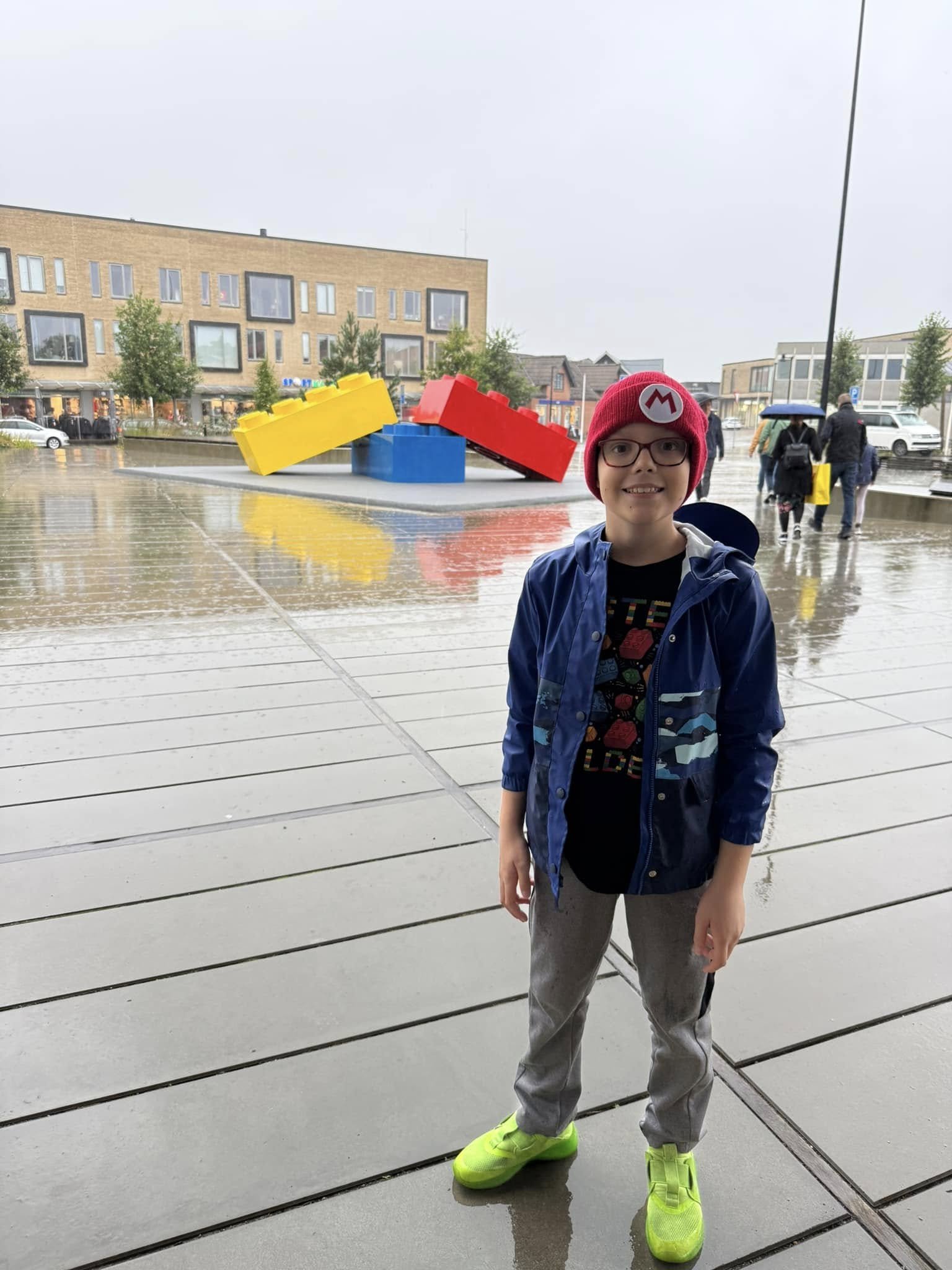 Young boy standing on wet pavement in front of a large colorful LEGO sculpture, wearing a red Mario hat, glasses, a blue rain jacket, gray pants, and bright yellow shoes, on a rainy day.