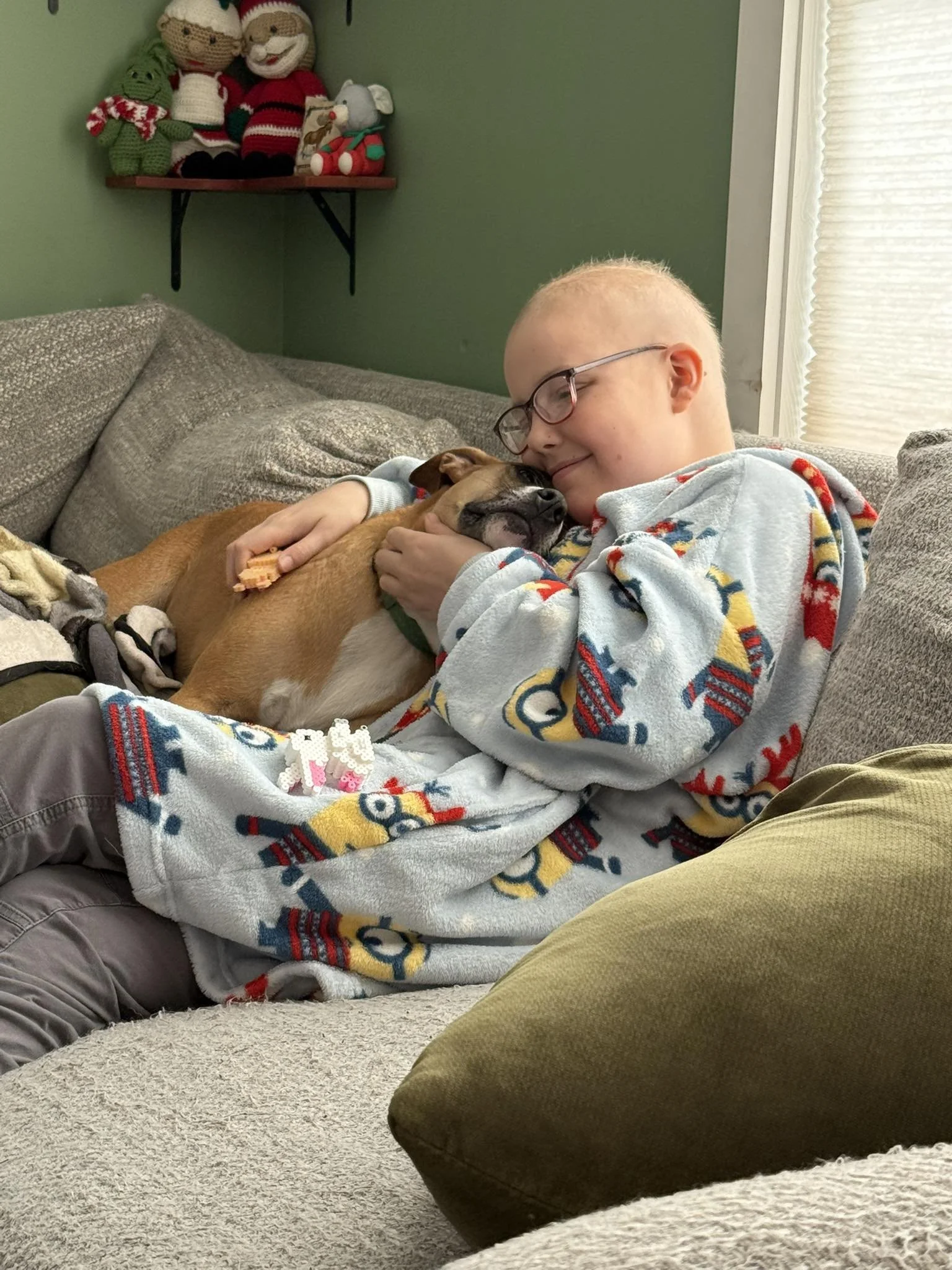 A young person wearing glasses and a holiday-themed blanket is cuddling with a tan and black dog on a gray couch in a living room, with holiday decorations on a shelf in the background.