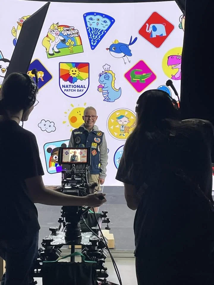 Boy standing in front of a colorful backdrop with cartoon characters and logos, being filmed or photographed by two people.
