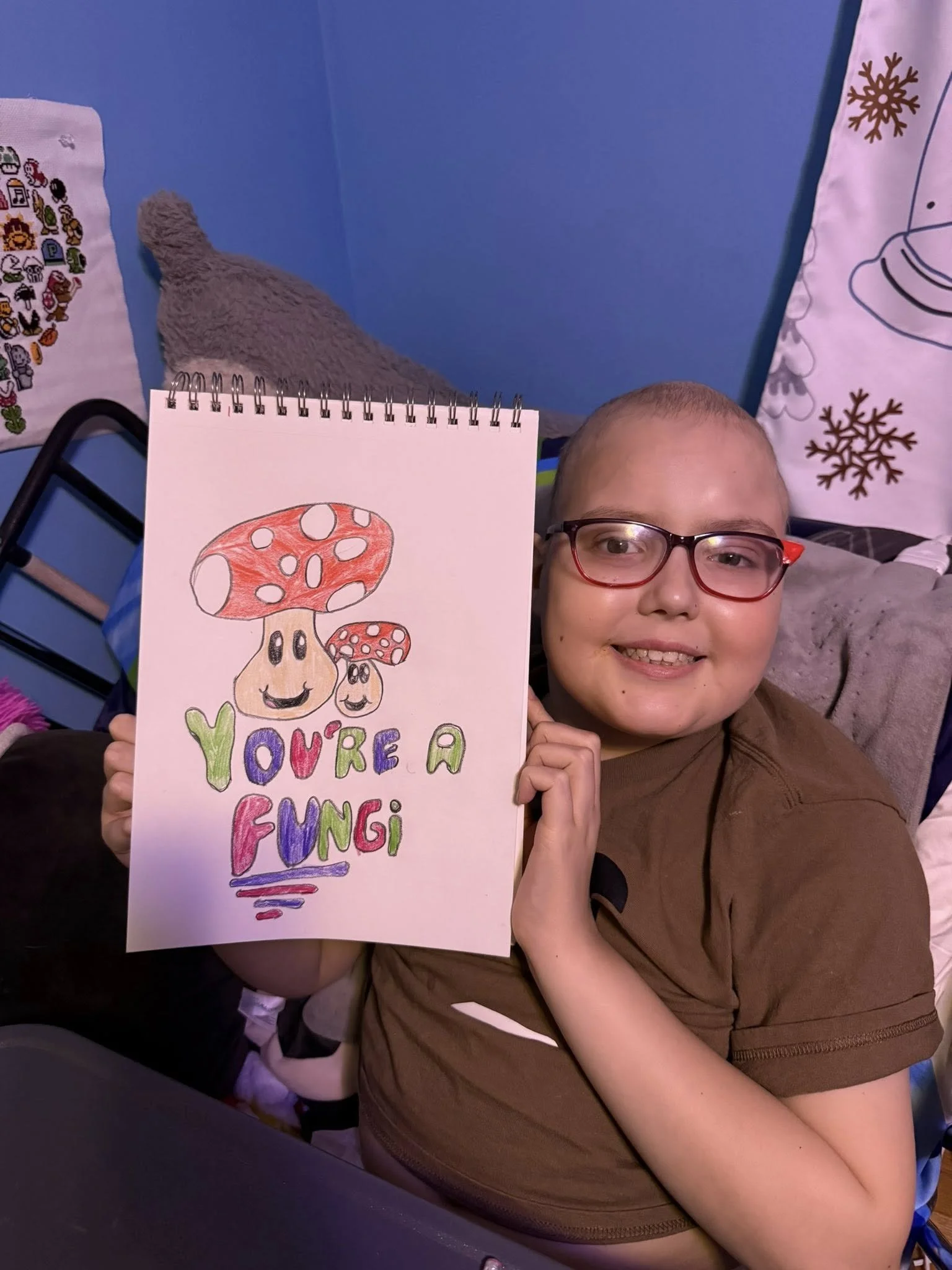 A smiling child with glasses holding a colorful drawing that says 'You're a Fungi' and features two animated mushrooms with smiling faces.
