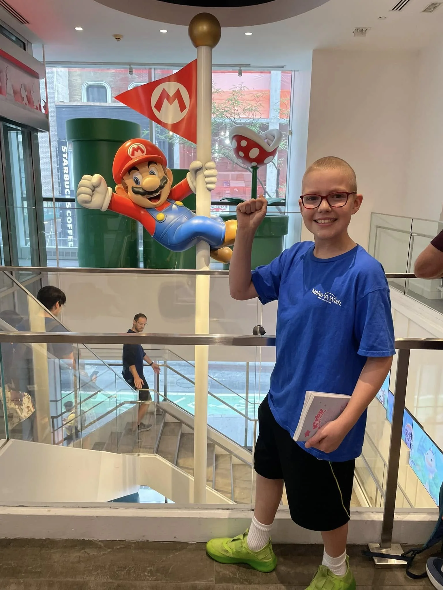 A young boy with glasses smiling and holding a Super Mario-themed flag and a mushroom toy at an indoor location with a large Mario statue in the background.