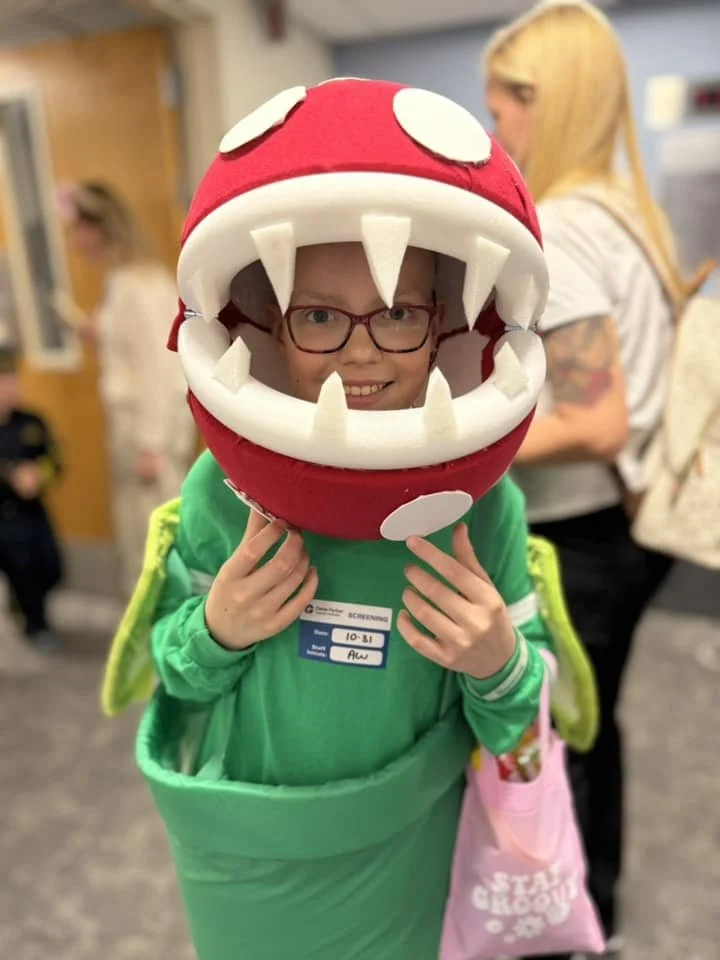Child in a green costume with a big red and white hat resembling a piranha plant from Mario, smiling through the plant's open mouth.