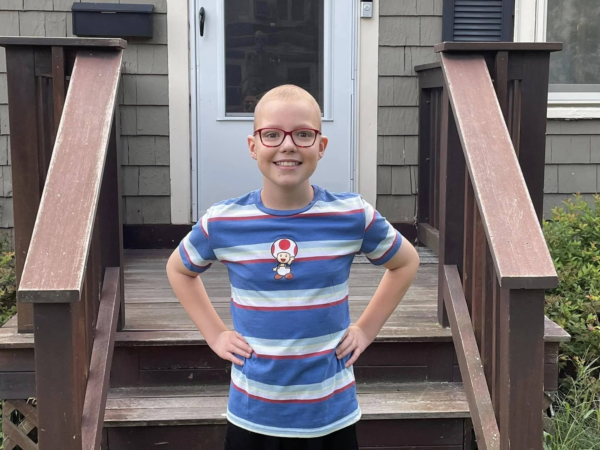 A smiling young girl with short hair and red glasses standing on a wooden porch with stairs, wearing a blue striped T-shirt with a Super Mario Toad character graphic, hands on hips, in front of a house entrance.