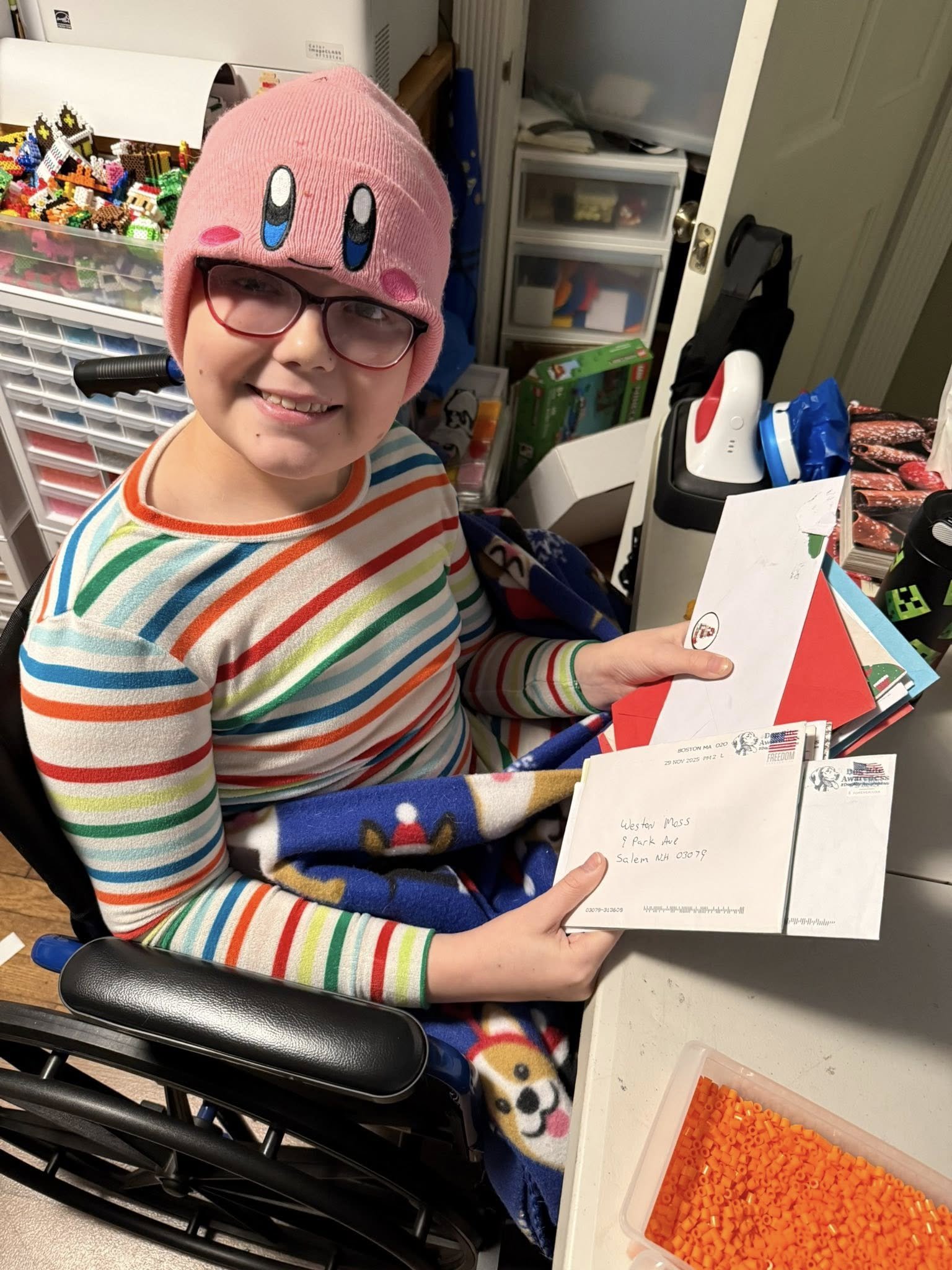 A smiling young person in a pink Kirby hat and rainbow striped shirt sitting in a wheelchair, holding envelopes, with a colorful blanket featuring cartoon characters, and surrounded by craft supplies like letters and beads.