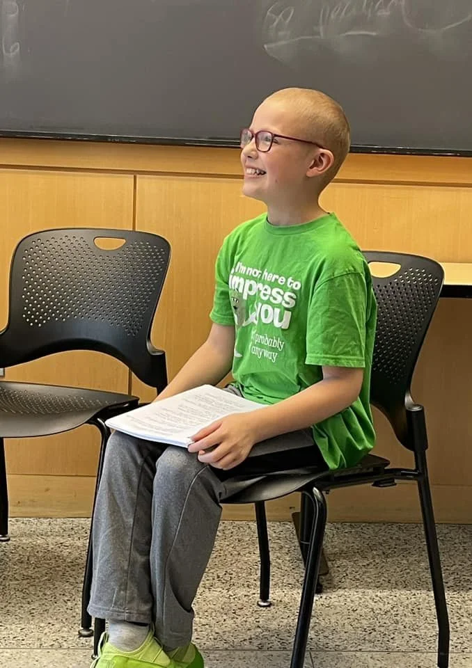 A young boy with glasses, wearing a bright green t-shirt and gray pants, sitting on a black chair in a classroom, smiling and holding a notebook.