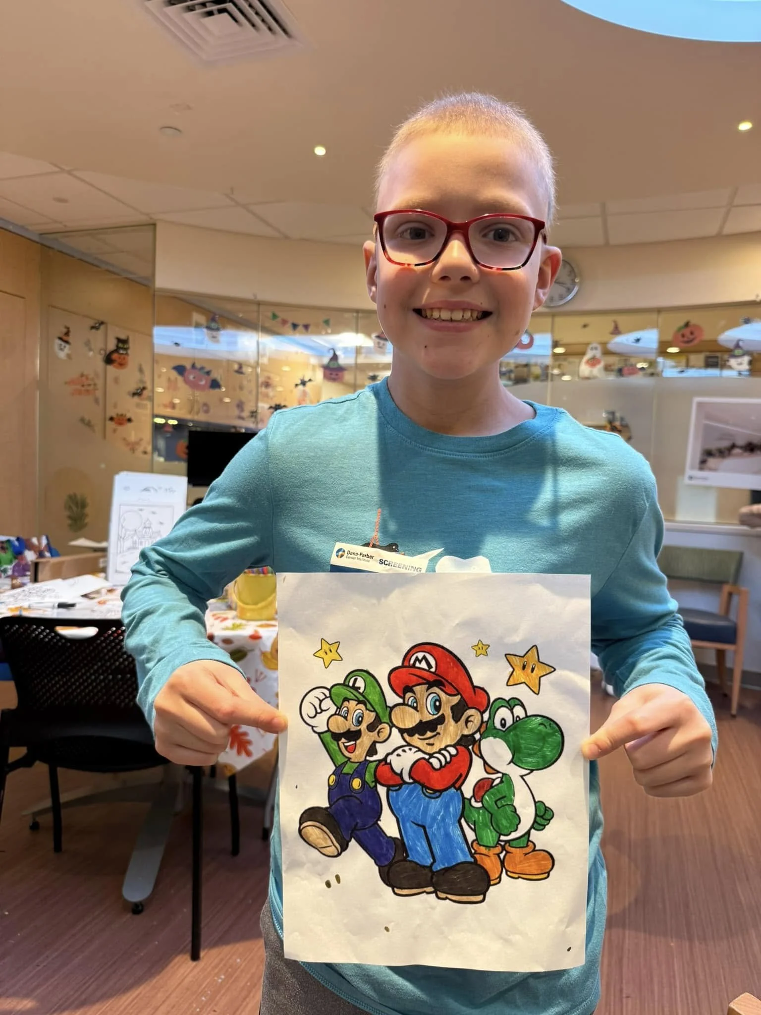 A smiling boy with glasses holding a drawing of Mario, Luigi, and Yoshi from Nintendo, pointing at the artwork with both hands.