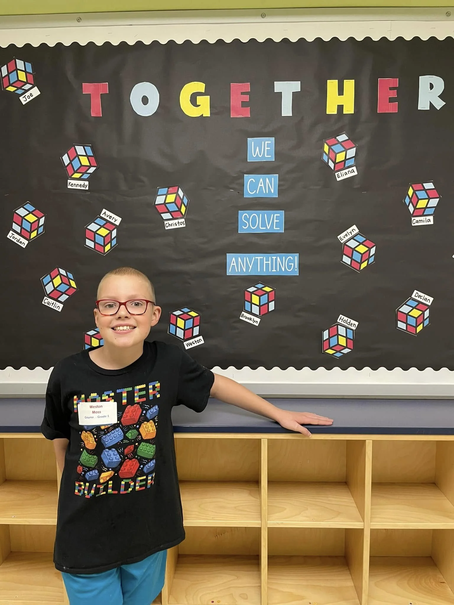 A young boy with red glasses and a black T-shirt stands in front of a classroom bulletin board. The board has the phrase 'Together We Can Solve Anything!' and colorful Rubik's Cube illustrations with students' names around it.