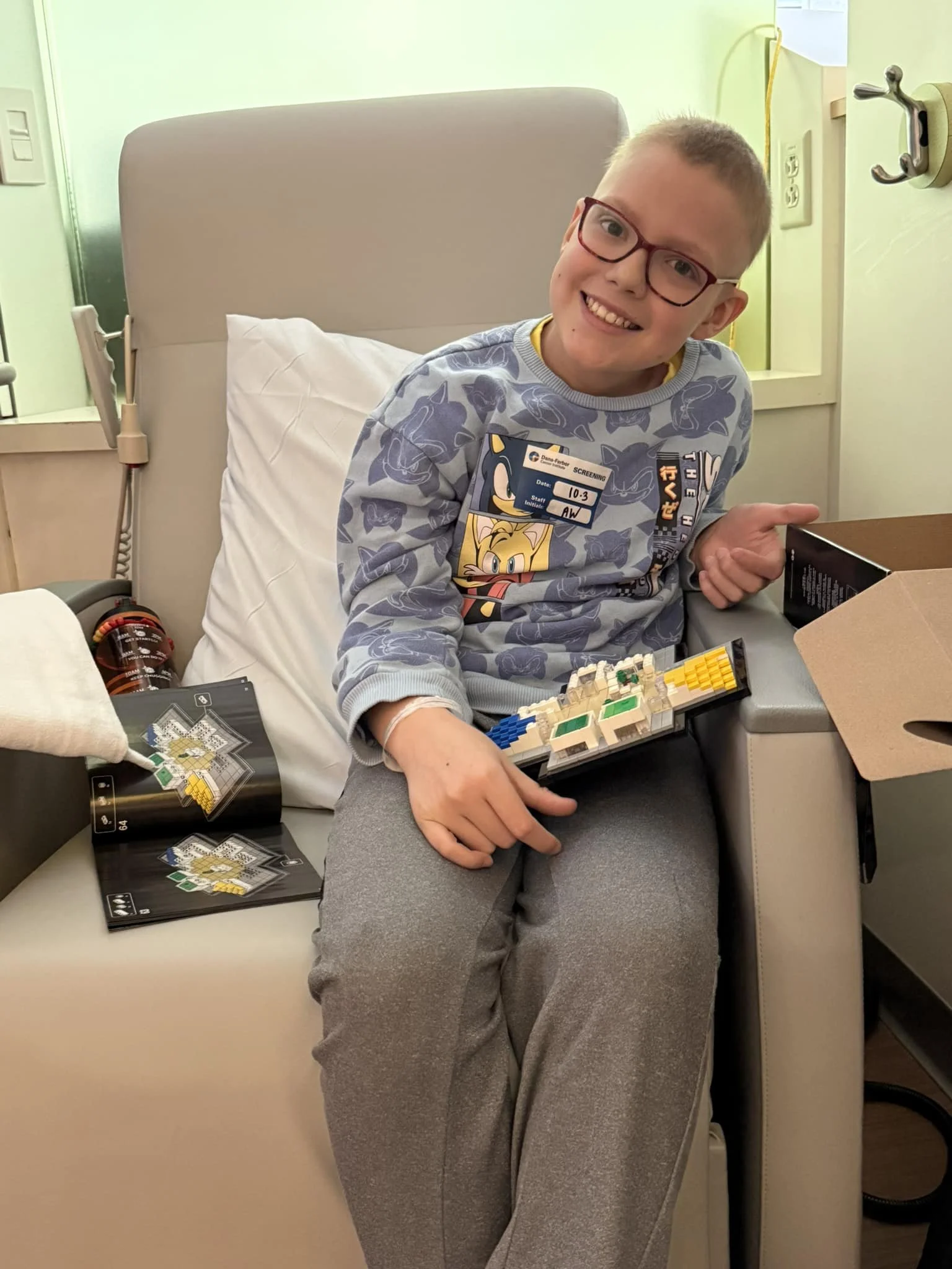 Young boy with glasses smiling, sitting in a hospital bed, holding a Lego set, with a cribside magazine open, hospital room with medical equipment and a window in the background.