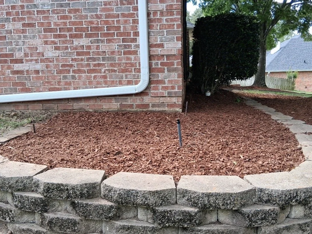 A landscaped area with red mulch, edged by a curved stone border, beside a brick house with a white gutter pipe, along a stone pathway, and a large green bush in the background.