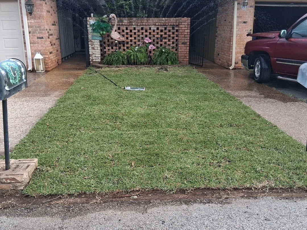 A front yard with freshly mowed grass, a mailbox on the left, a brick house with a decorative pink flamingo statue and plants, a red pickup truck parked on the driveway, and a sprinkler placed in the grass.