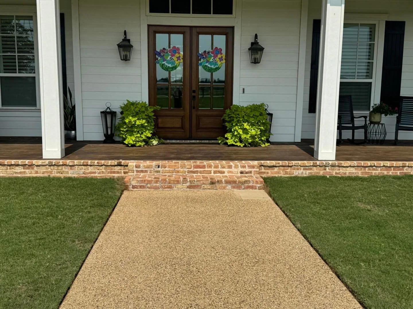Front porch of a house with brick steps leading to a wooden door with glass panels, decorated with colorful Easter-themed window decals. The porch has two large green bushes, black lanterns, and seating, with grass and a sidewalk in the foreground.