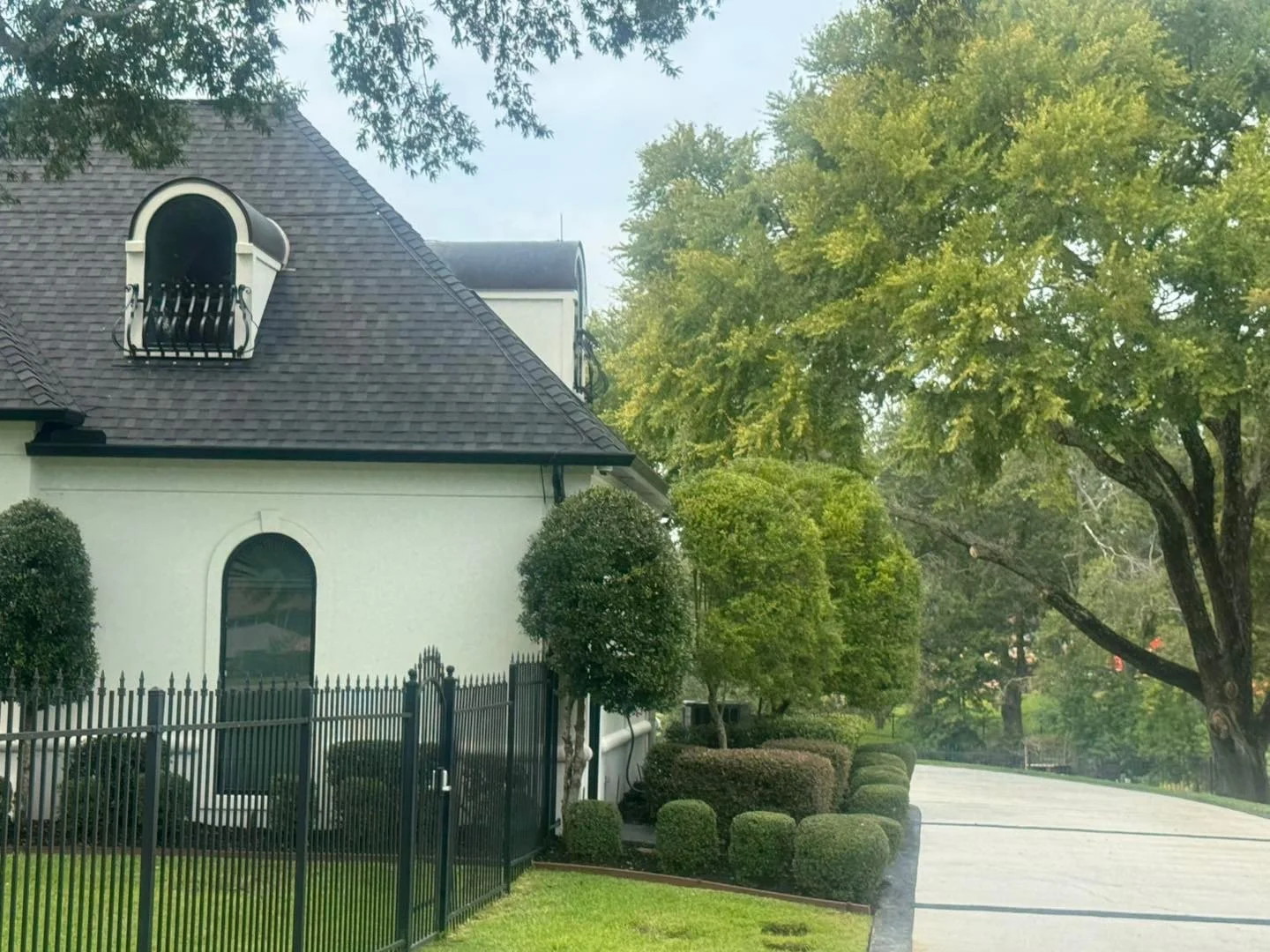 A white building with a steep roof, black fence, and manicured bushes, surrounded by green trees, during daytime.