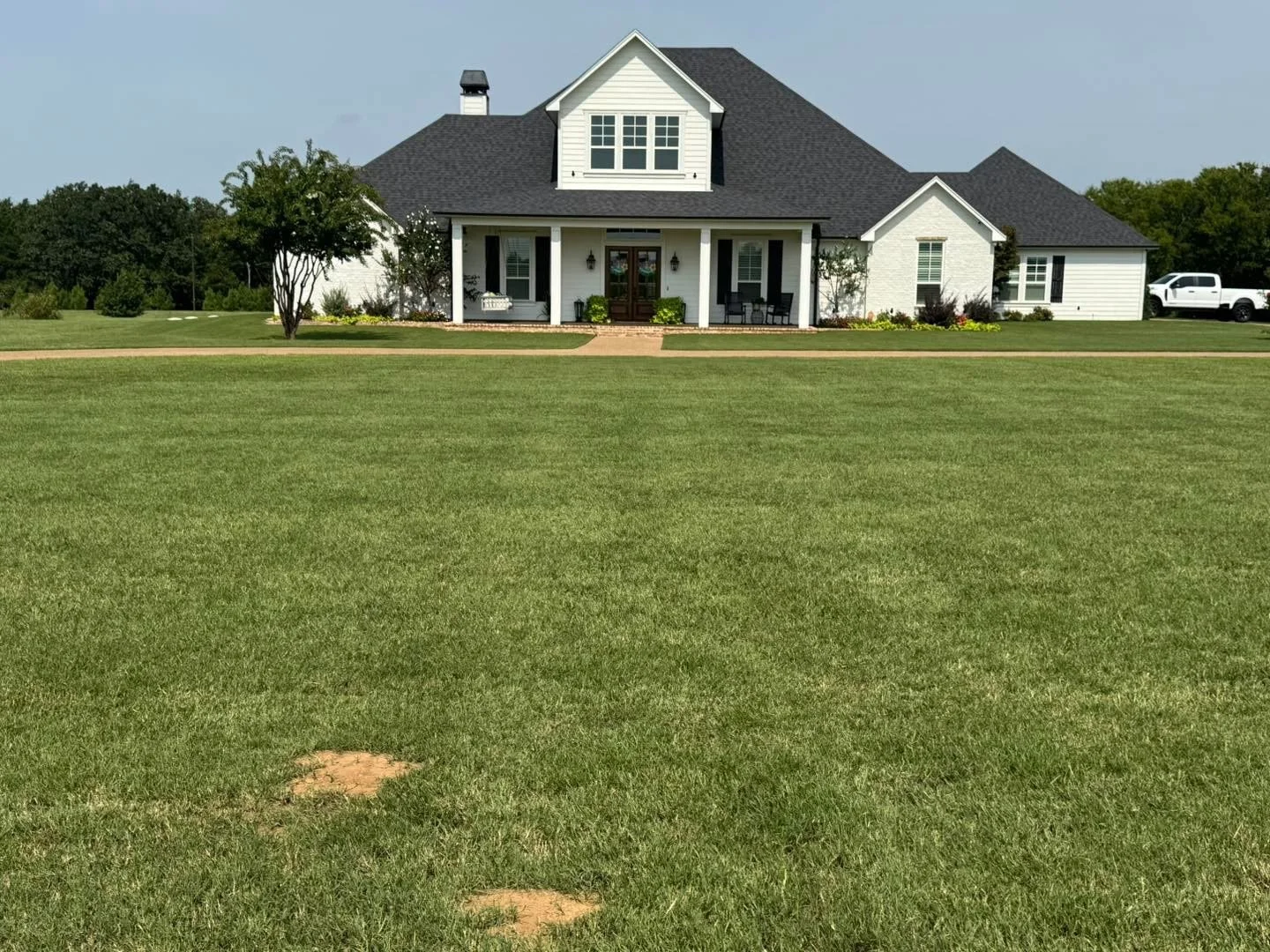 Front view of a large white house with a dark gray roof and a porch, surrounded by a well-maintained lawn and trees, under a blue sky.