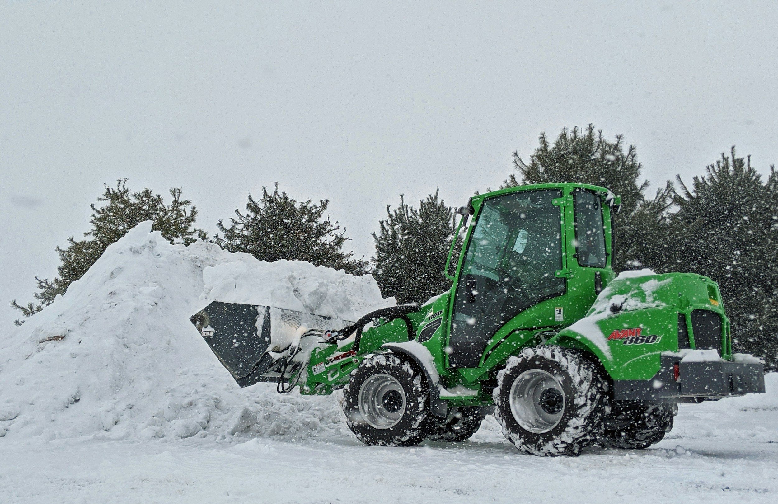 Green compact loader clearing snow in an outdoor area with pine trees and snowfall.