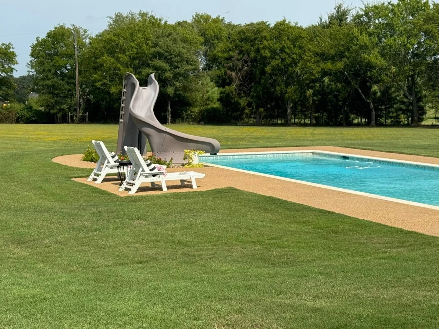 A backyard scene with a swimming pool, a gray water slide, two white lounge chairs, and a small round table with flowers, surrounded by green grass and trees in the background.