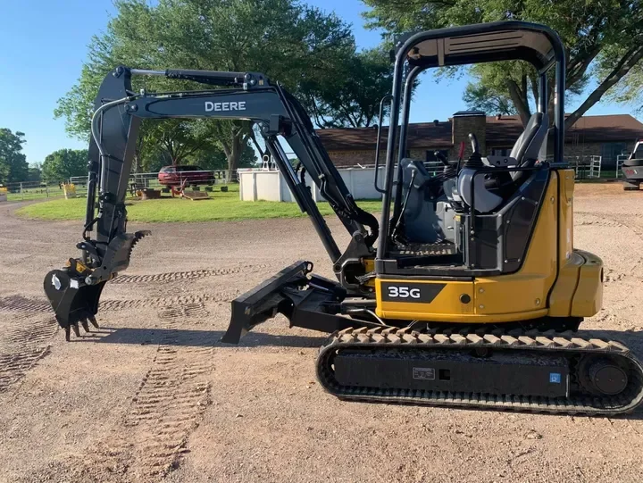 Yellow and black compact excavator on a dirt surface with trees and a building in the background.