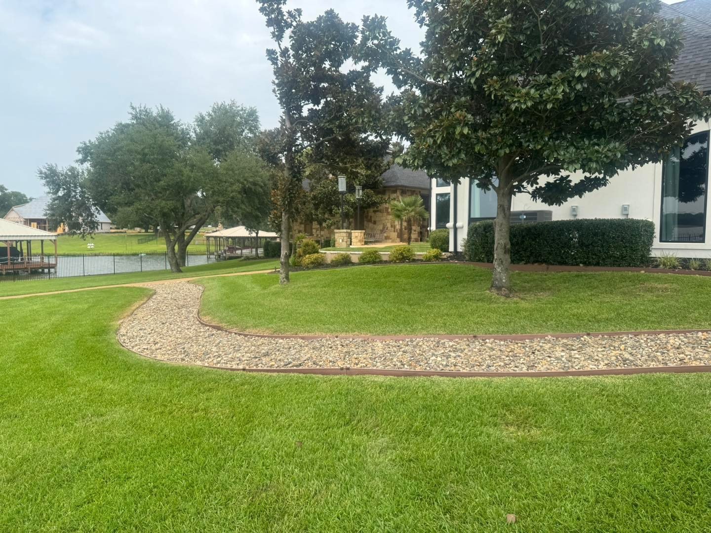 A well-maintained backyard with green lawn, a curved gravel pathway, and several trees, with a neighboring house and body of water in the background.