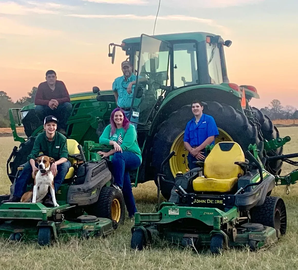 Group of five people and a dog posing in front of a large green John Deere tractor on a grassy field at sunset.