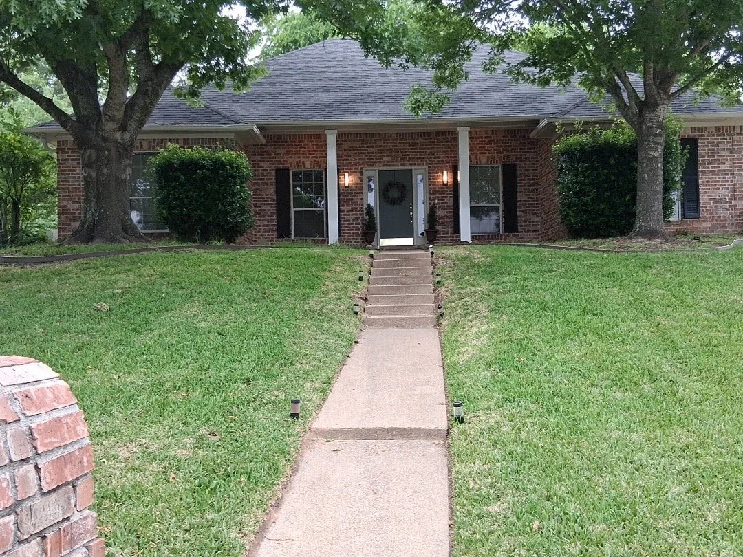 Front view of a brick house with a dark front door decorated with a wreath, flanked by two potted plants, and a concrete walkway leading up stairs to the entrance. There are trees on both sides of the house and neatly trimmed bushes near the windows.