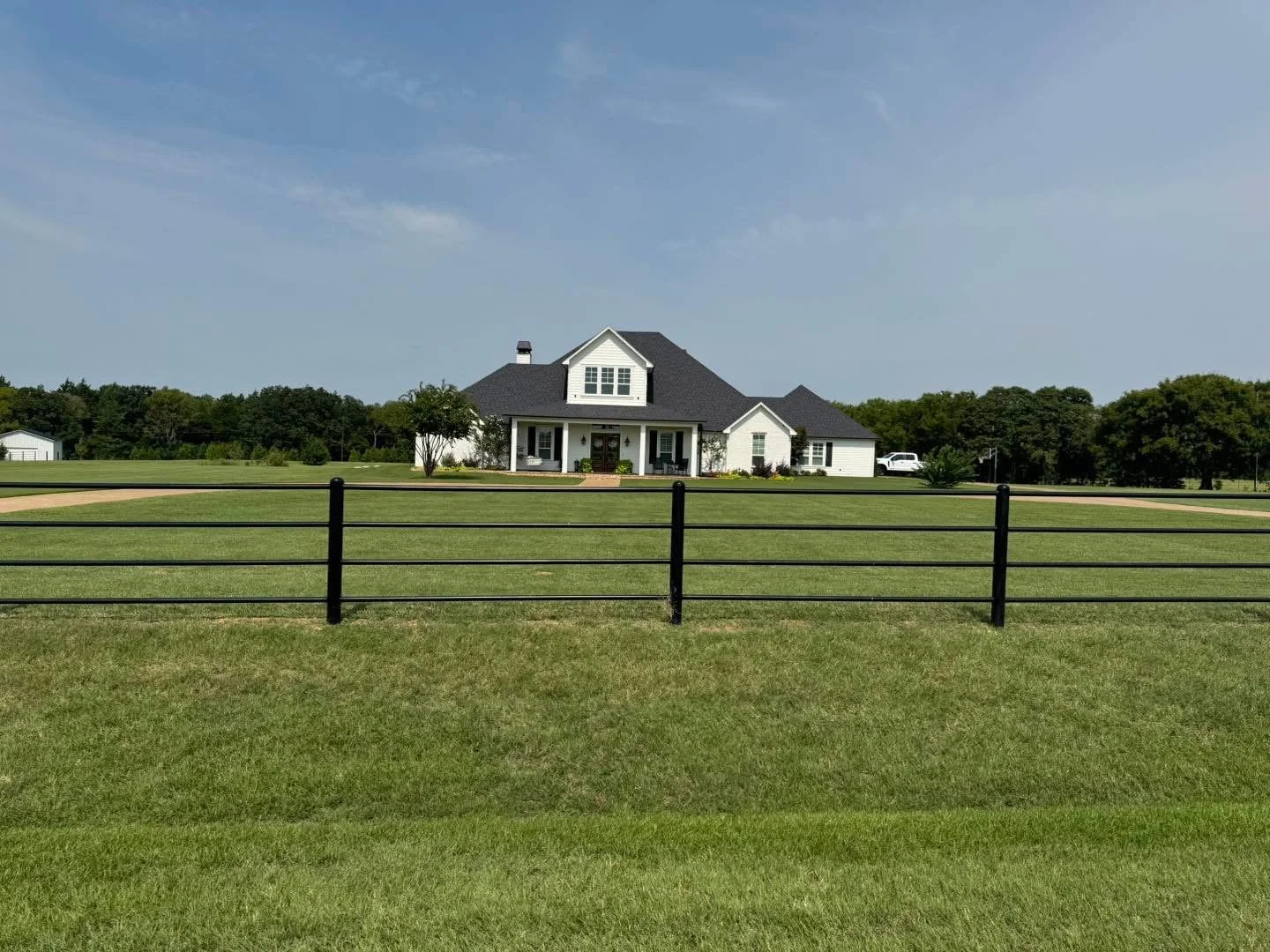 Large white house with black roof, surrounded by green lawn and trees, view from a fenced yard under a blue sky.
