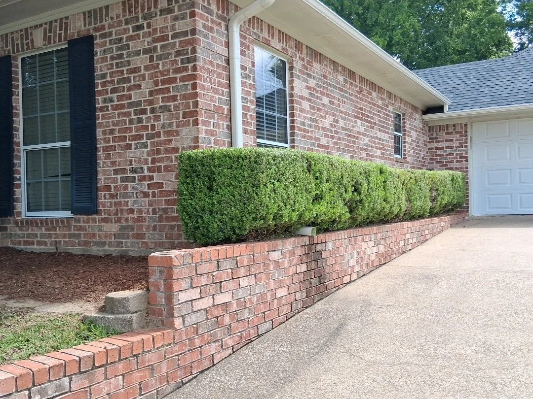 A brick house with black shutters and a white garage door. Well-maintained green bushes in front of the house and a brick retaining wall separating the bushes from the driveway.