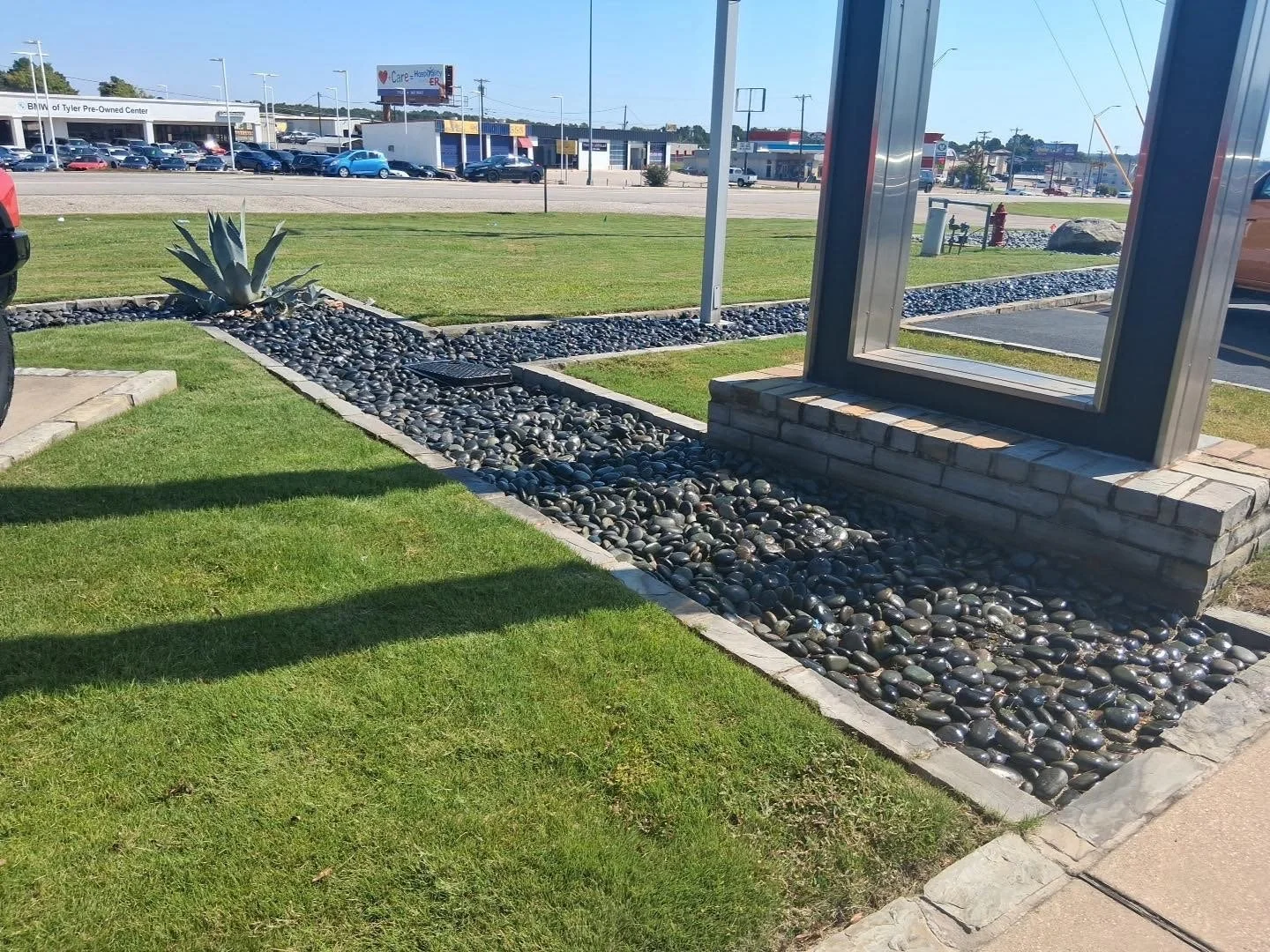 A small landscaped area with a Agave plant and black river rocks, bordered by bricks, next to a large parking lot and a street with cars, billboards, and shops in the background.