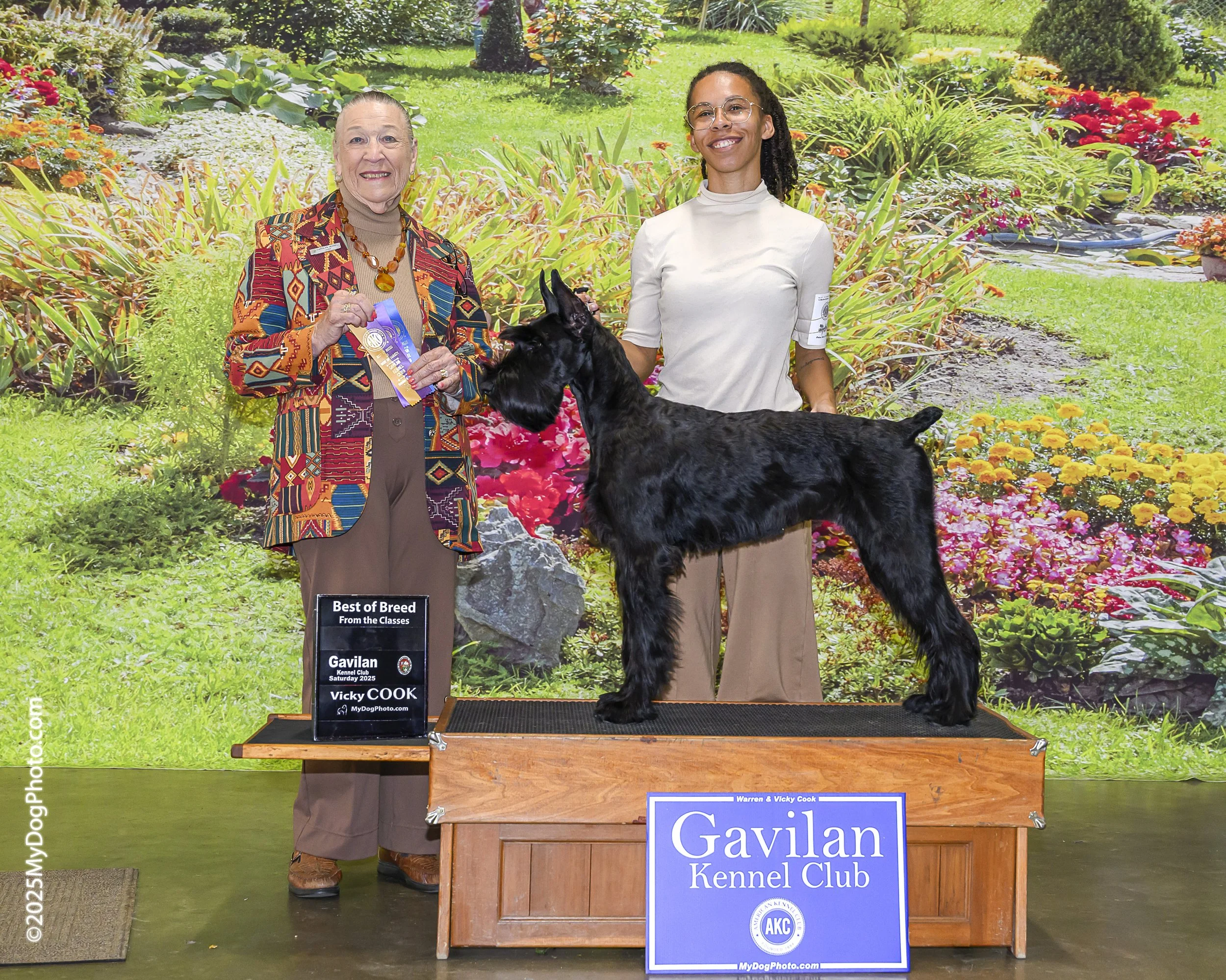 A dog show award ceremony at Gavilán Kennel Club. Two women stand behind a black Scottish Terrier dog on a wooden platform. The woman on the left is smiling and holding a ribbon, wearing a colorful patterned blazer. The woman on the right wears glasses and a beige top, smiling as well. The background displays a garden scene with flowers and green plants.