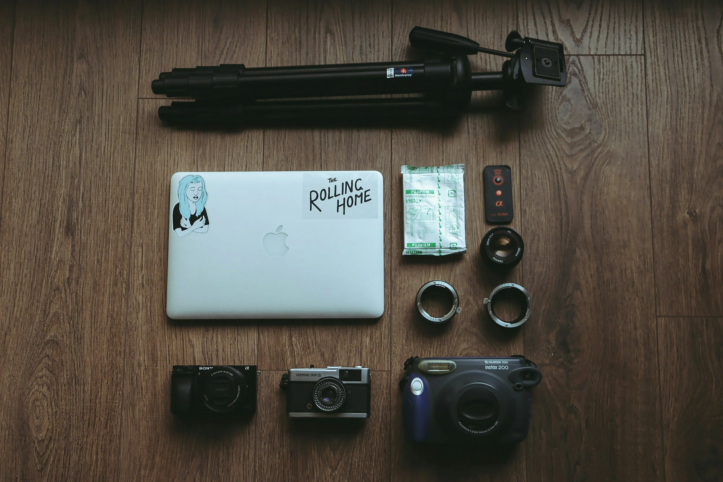 A collection of photography and filming equipment on a wooden floor, including a tripod, a closed MacBook with a sticker that says "The Douling Home," a packet of instant coffee, a remote control, two camera lens adapters, two cameras (Sony compact camera and vintage film camera), and a large waterproof camera housing.