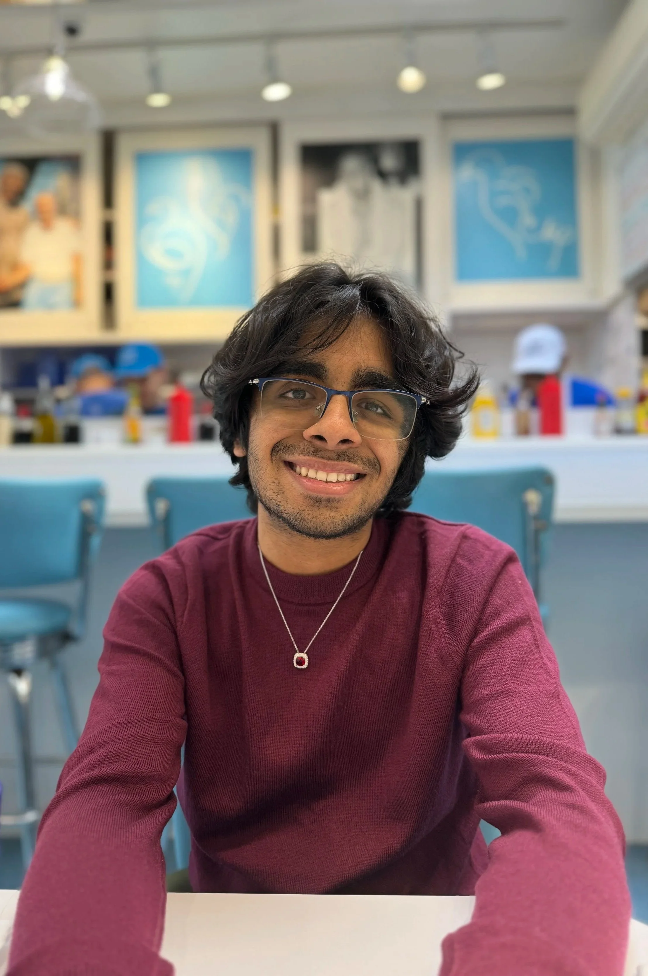 A young man with glasses and dark hair smiling at the camera, sitting at a table in a diner or cafe with blue chairs and a colorful background.