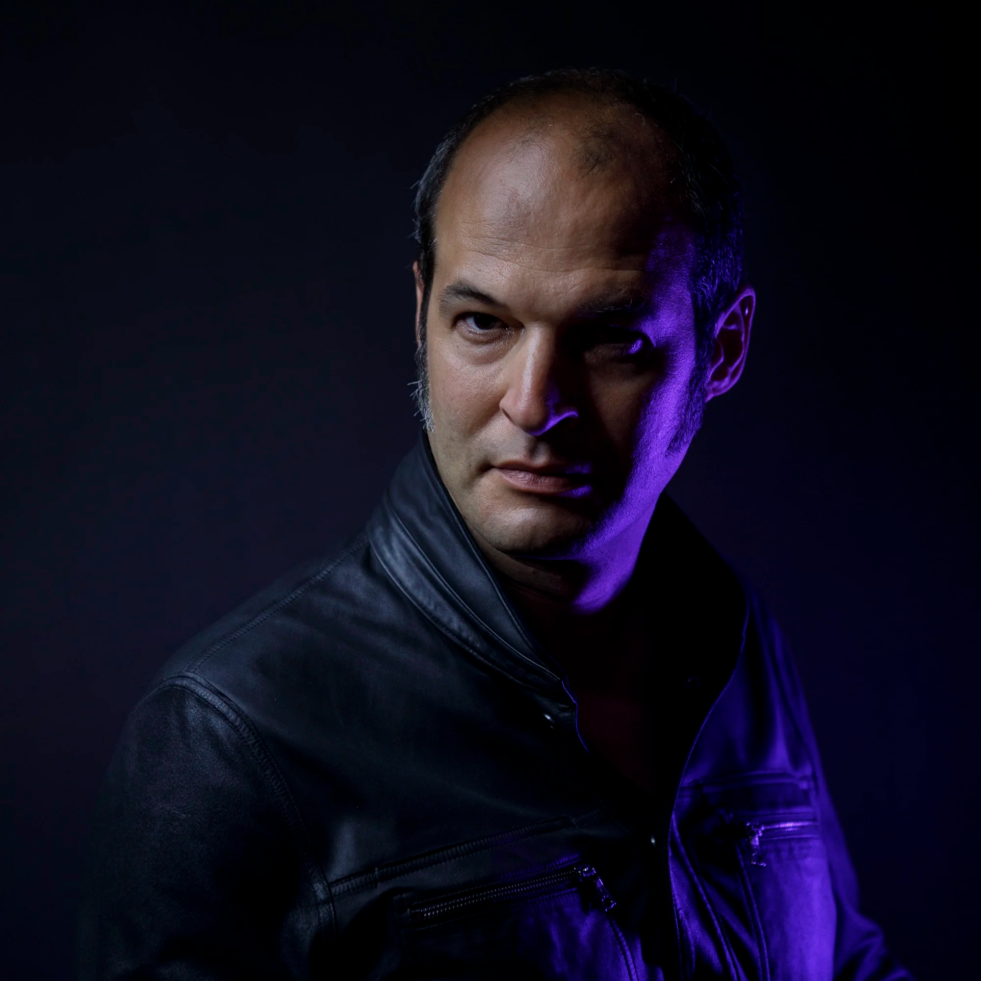 Portrait of Jez Harris in a leather jacket, looking sideways with serious expression, illuminated by purple and blue lighting.