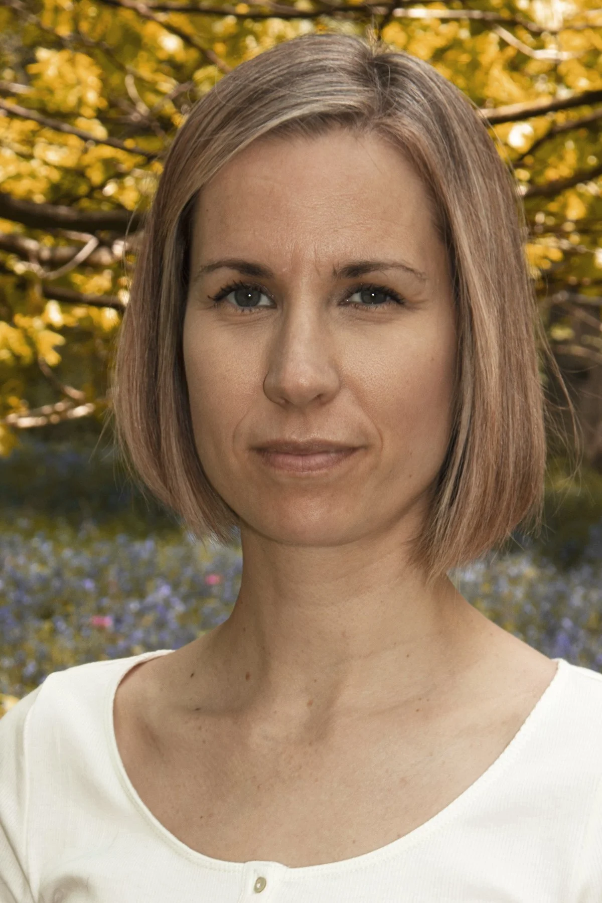 Portrait of a woman with short, blonde hair, wearing a white top, standing outdoors with yellow autumn leaves in the background.