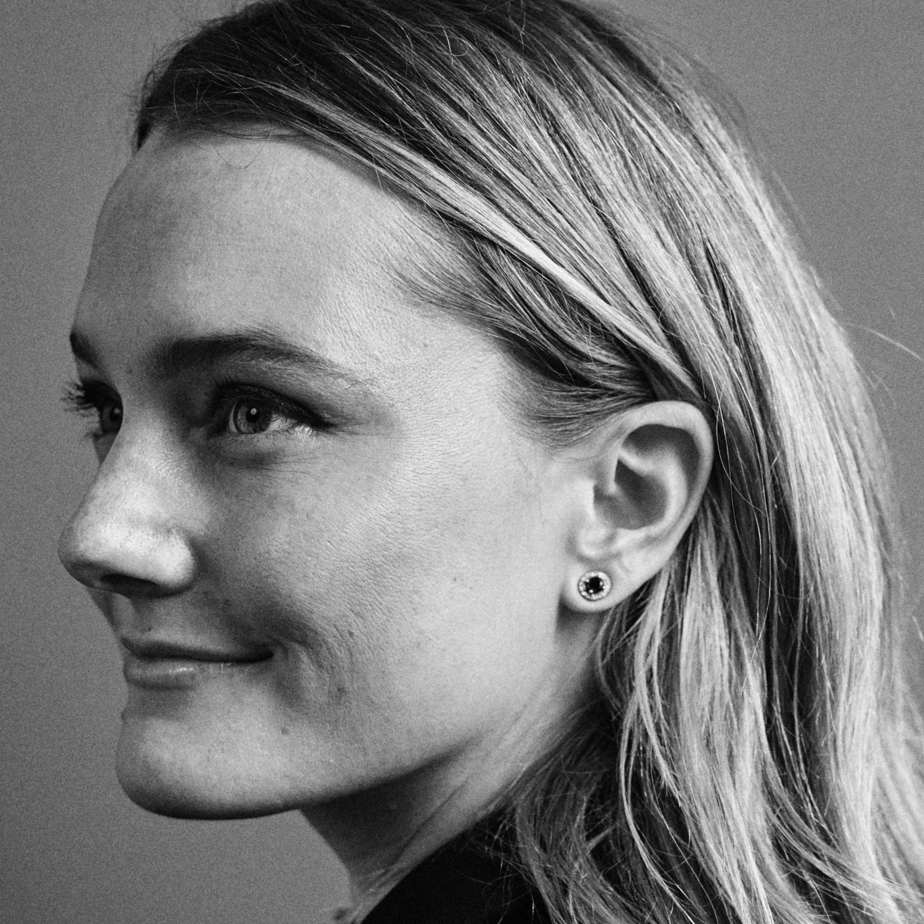 A black and white close-up portrait of a woman with light-colored hair, smiling slightly, wearing a small earring, looking to her left.