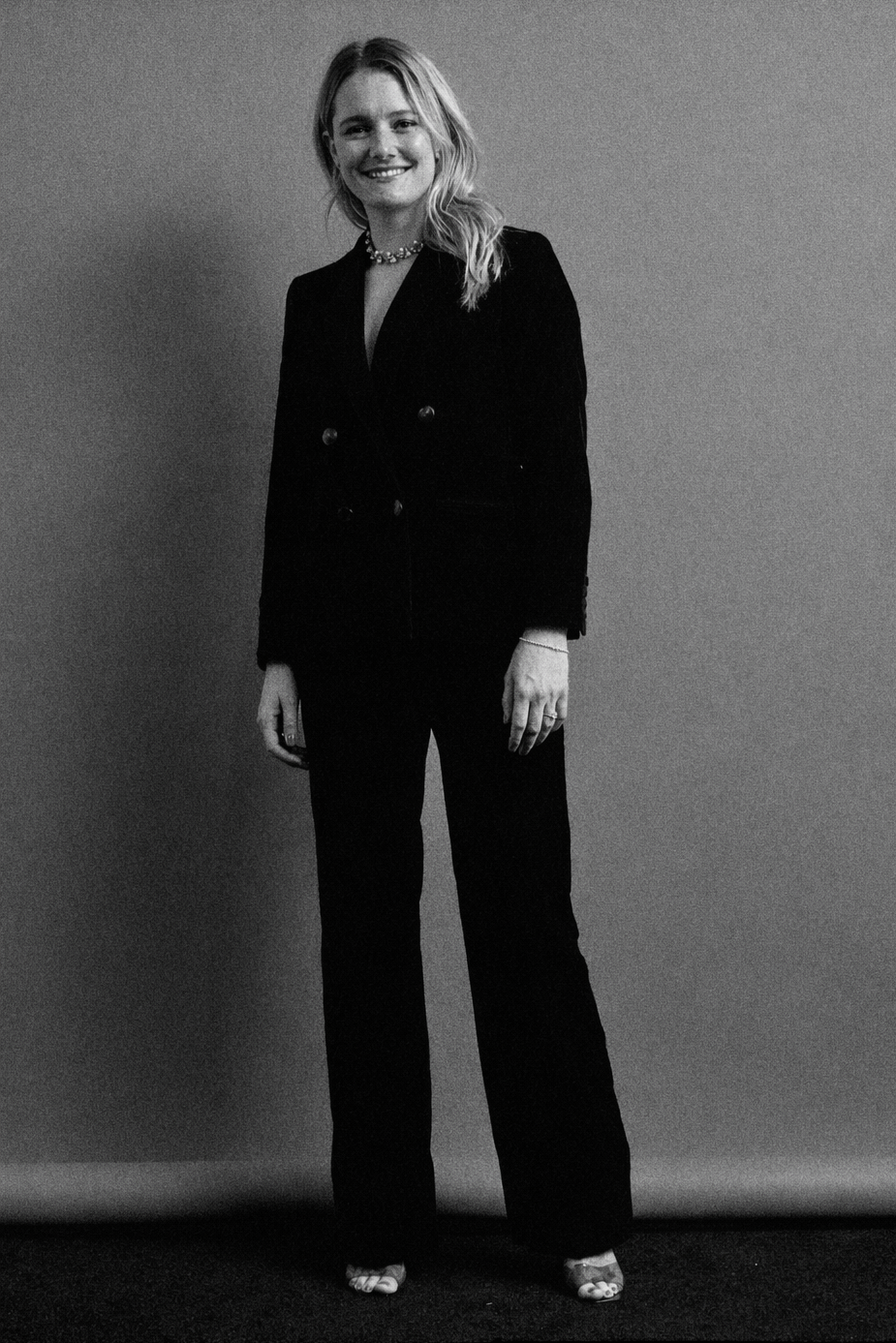 A woman standing against a plain wall, smiling and dressed in a black suit with a double-breasted blazer and matching trousers, wearing open-toed heels and a necklace.