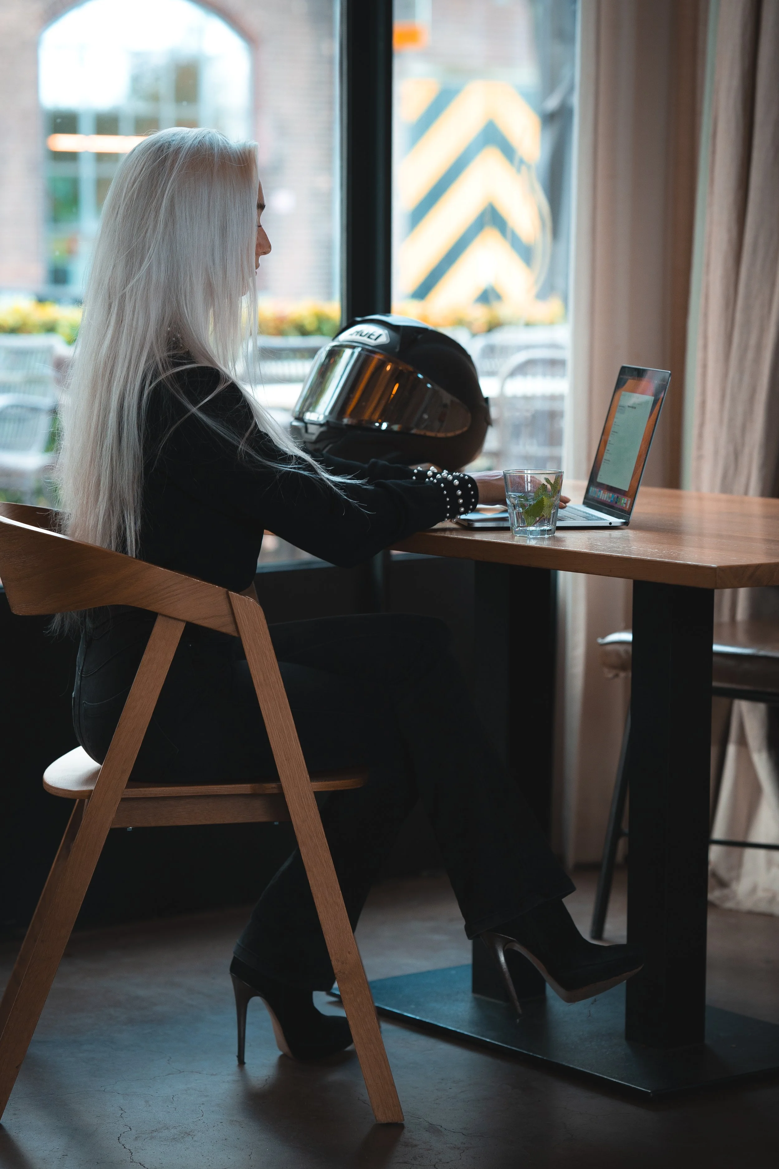 Een vrouw met lang blond haar zit aan een houten tafel in een café, werkt op haar laptop en heeft een helm naast haar staan.
