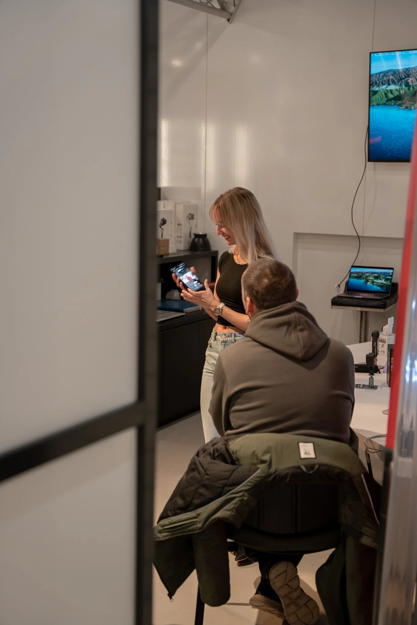 Een lachende vrouw die met een smartphone in haar hand staat in een technologiewinkel, terwijl een man met een hoodie op haar zit. Er zijn monitoren en laptops zichtbaar op de achtergrond.