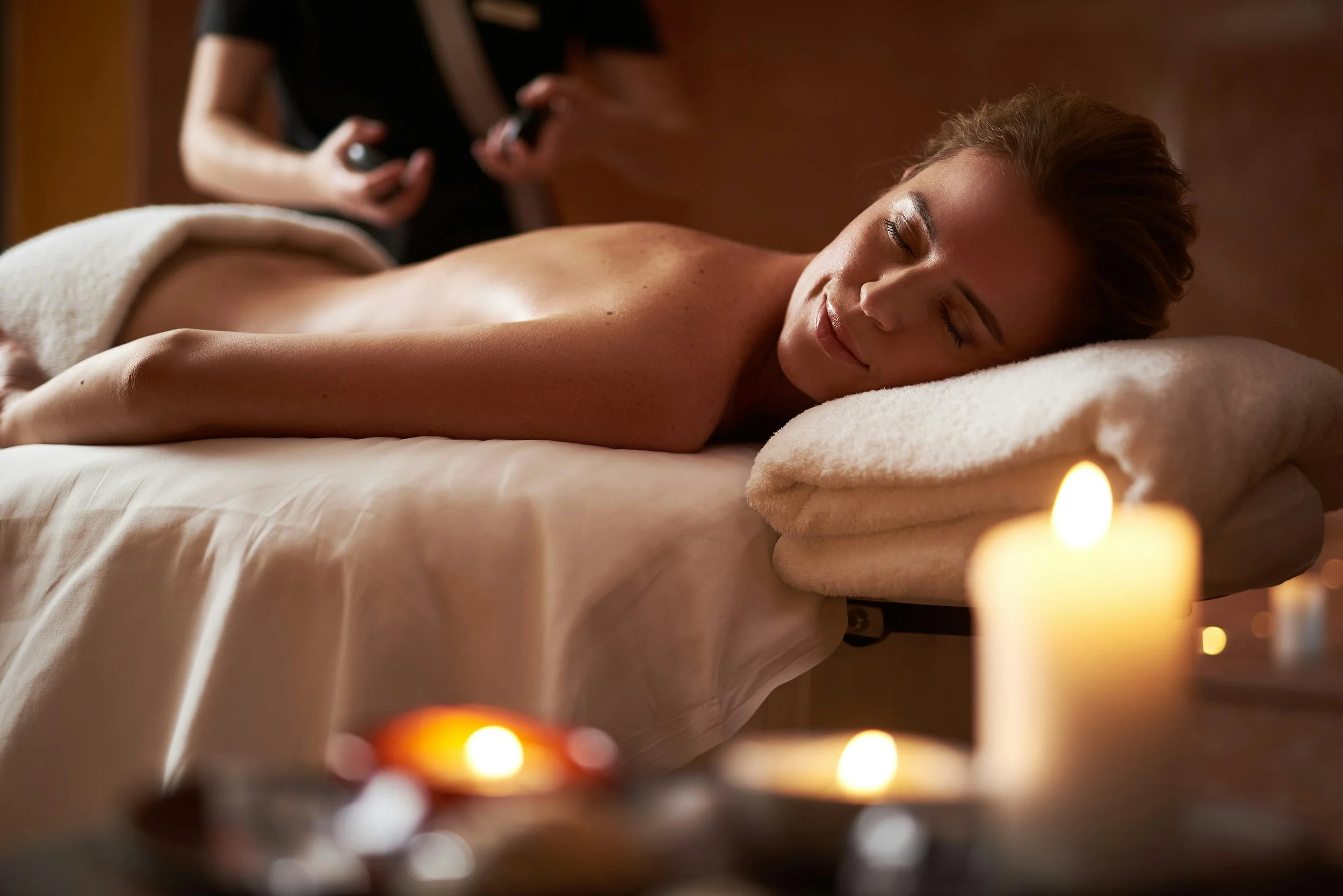 A woman lies face down on a massage table with her eyes closed, receiving a massage, with candles burning nearby creating a relaxing atmosphere.