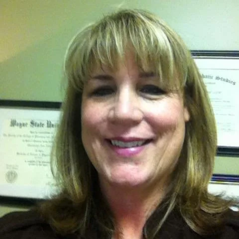Gwen Nelson smiling standing in front of a wall of her framed certificates or diplomas.