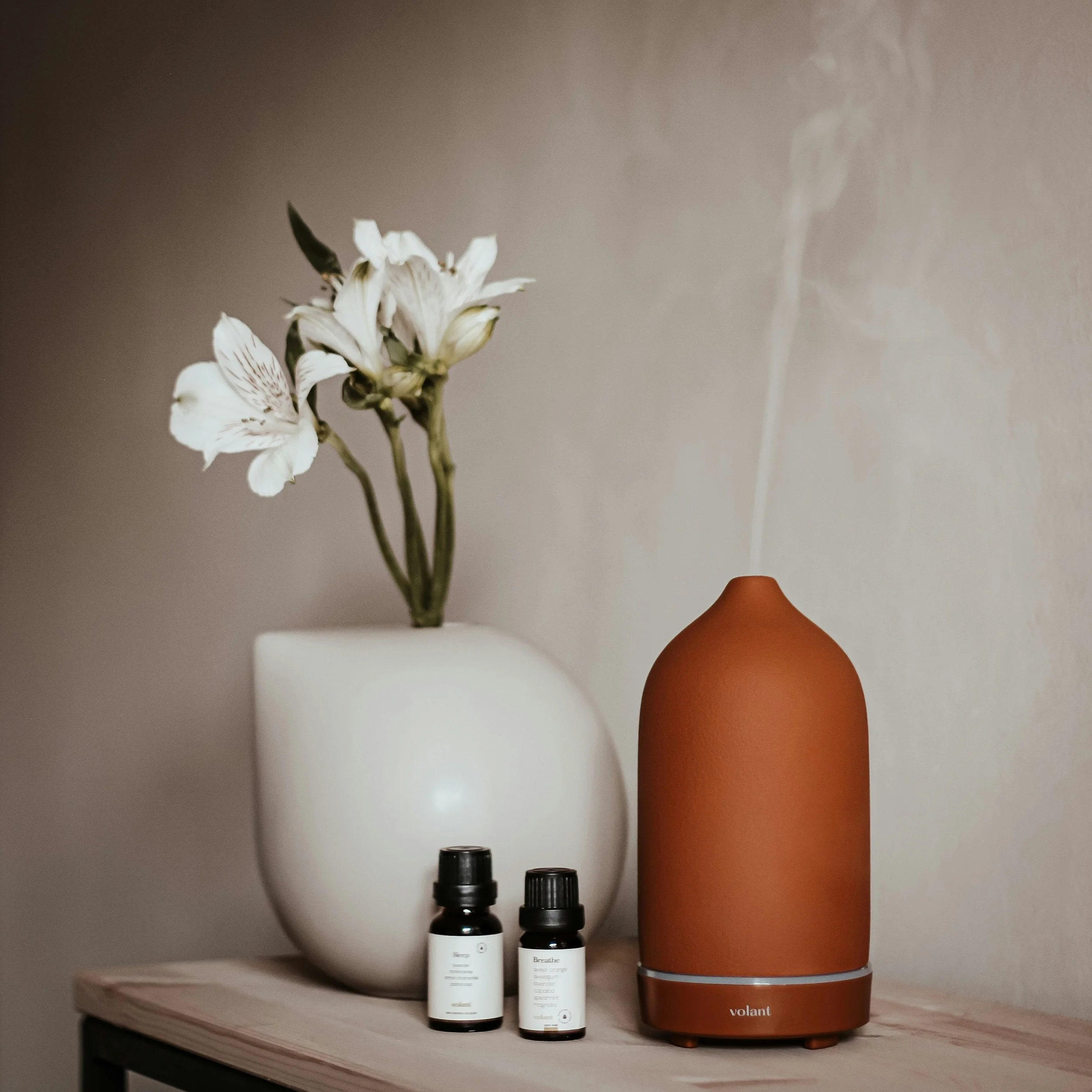 A white vase with white flowers, two small black bottles, and a rose-colored essential oil diffuser on a wooden surface against a neutral wall.