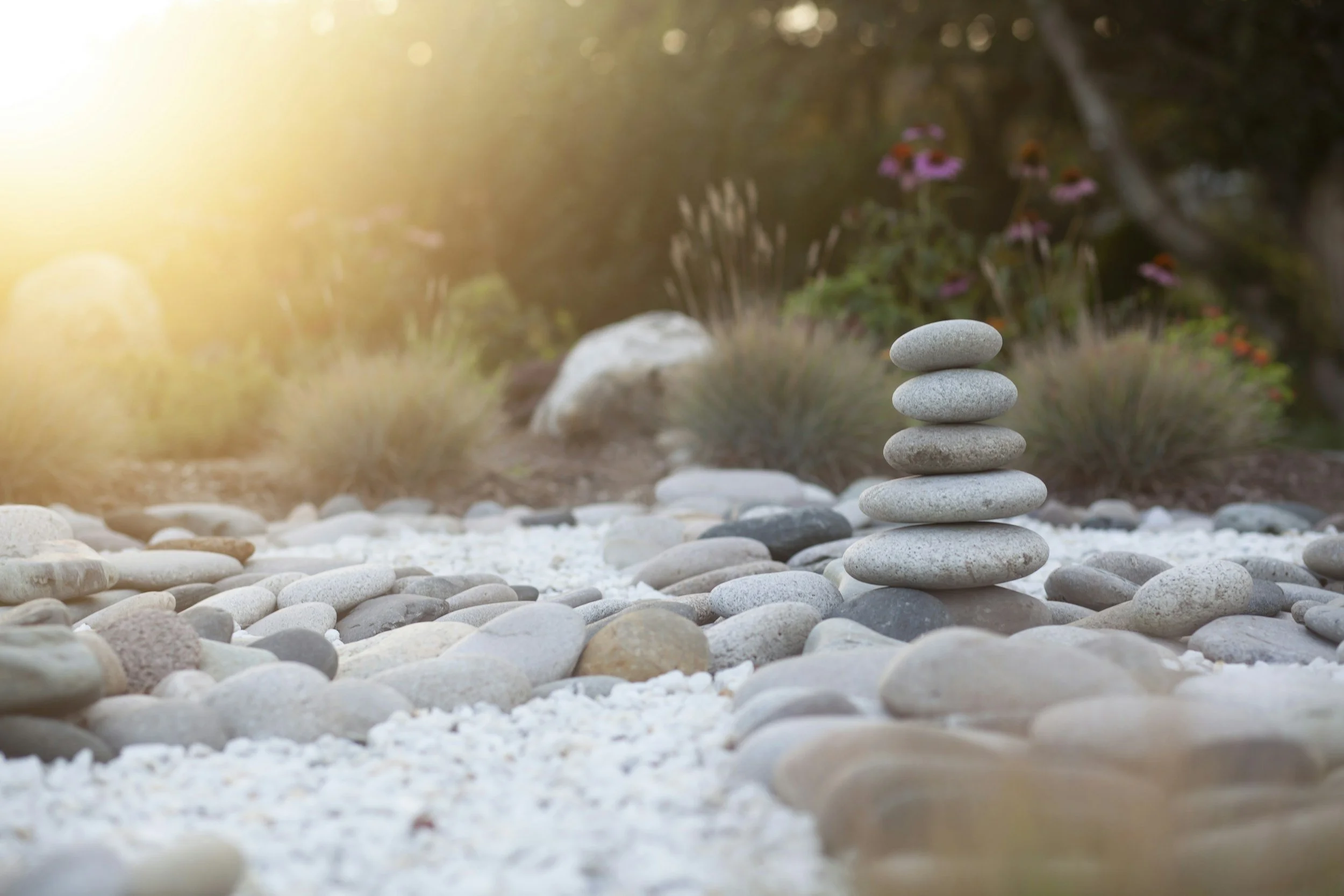 A zen garden with a stack of seven smooth gray stones in the foreground, surrounded by smaller rounded stones and gravel, with ornamental plants and flowers in the background, bathed in warm sunlight.