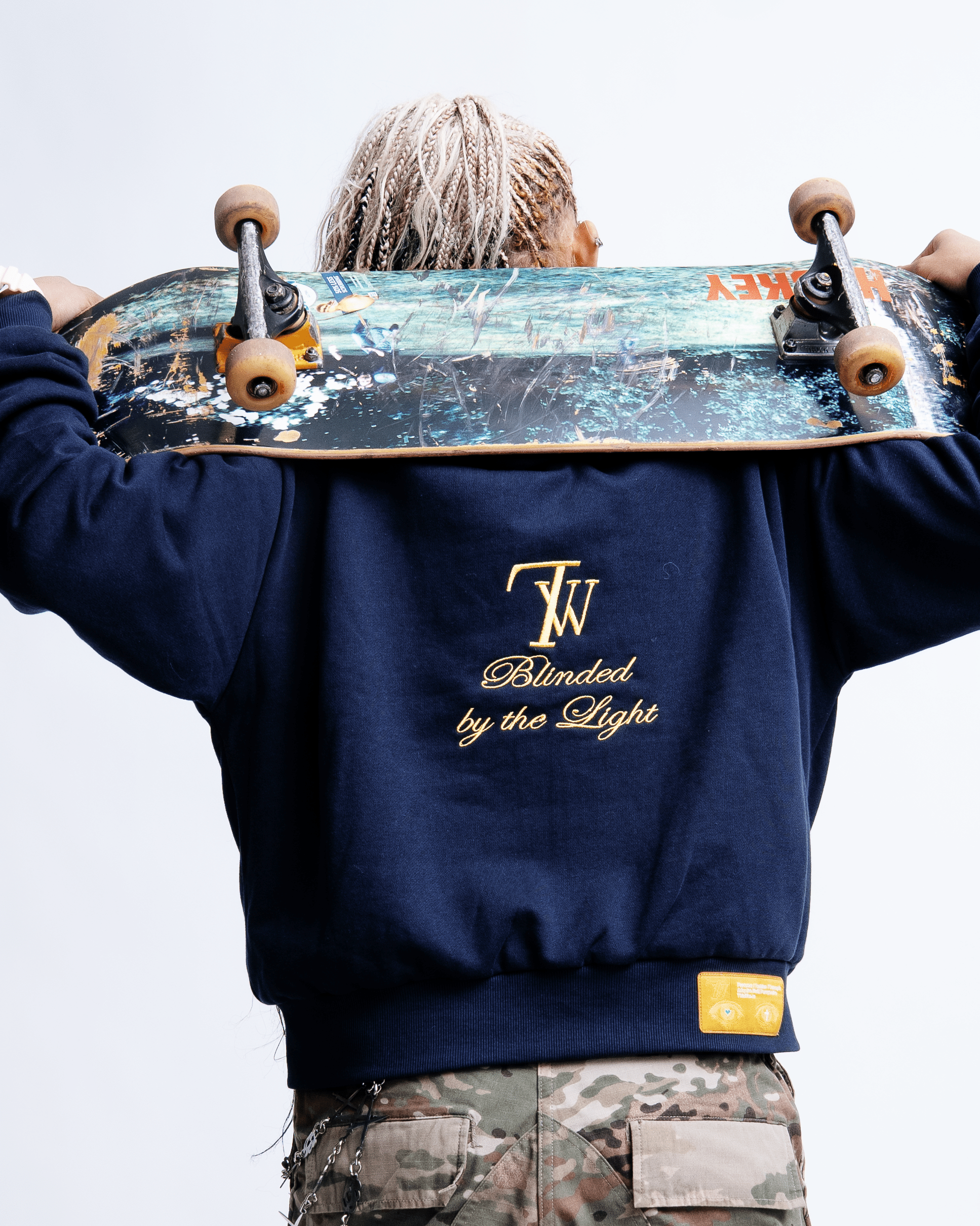 Person with dreadlocks holding a skateboard over their shoulders, wearing a navy sweatshirt with embroidered text and camouflage pants.