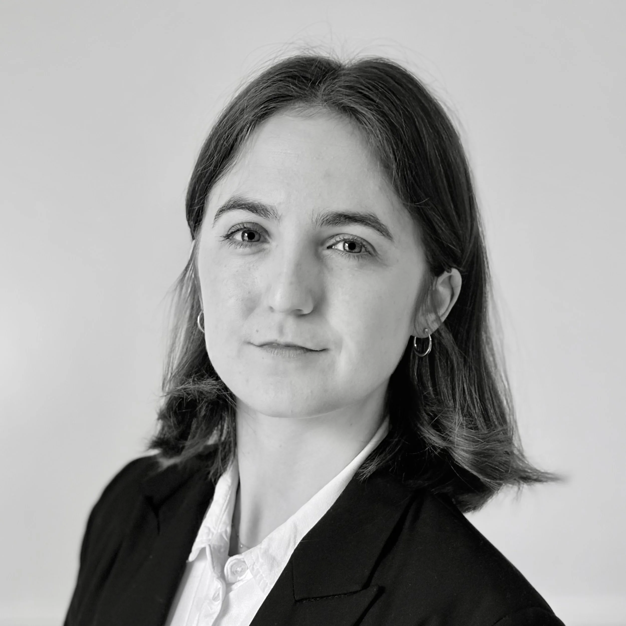Black and white photo of a young woman outdoors in a wooded area, wearing a jacket and smiling.