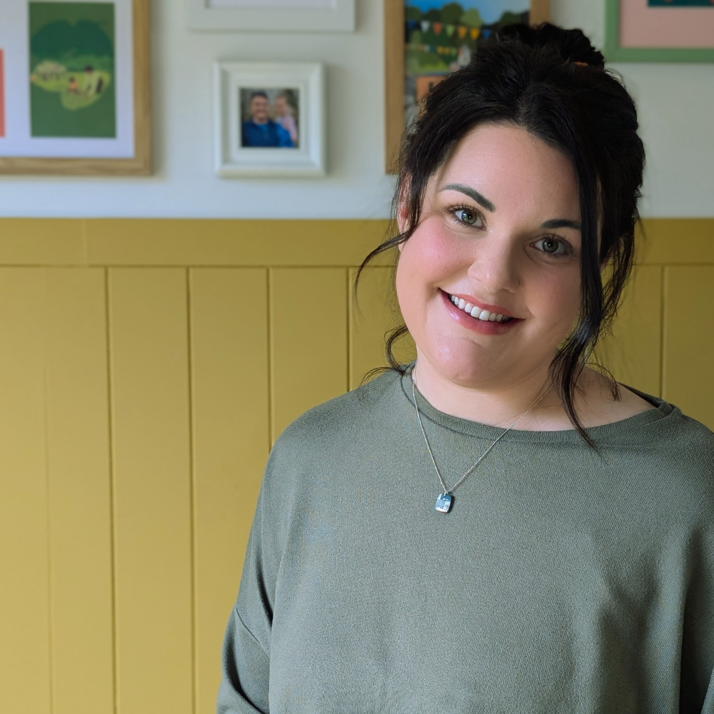 A smiling woman with dark hair and a green top standing indoors in front of a wall with framed photos and artwork.