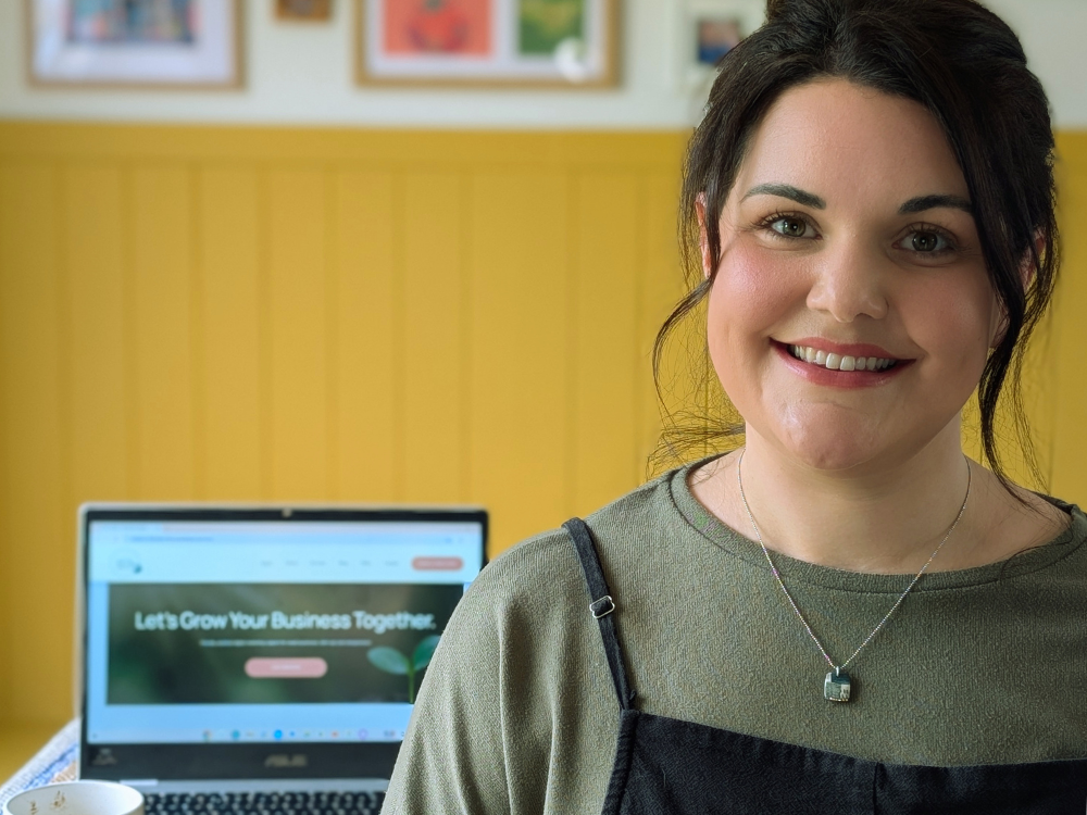 A woman smiling in a room with a yellow wall and photographs, with a laptop displaying a website that reads 'Let's Grow Your Business Together.'