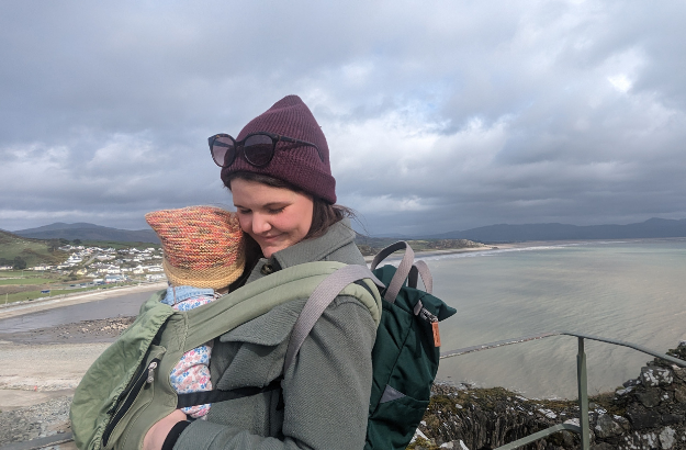 A woman holding a baby in a baby carrier outdoors near the ocean, with a rocky cliff and cloudy sky in the background.